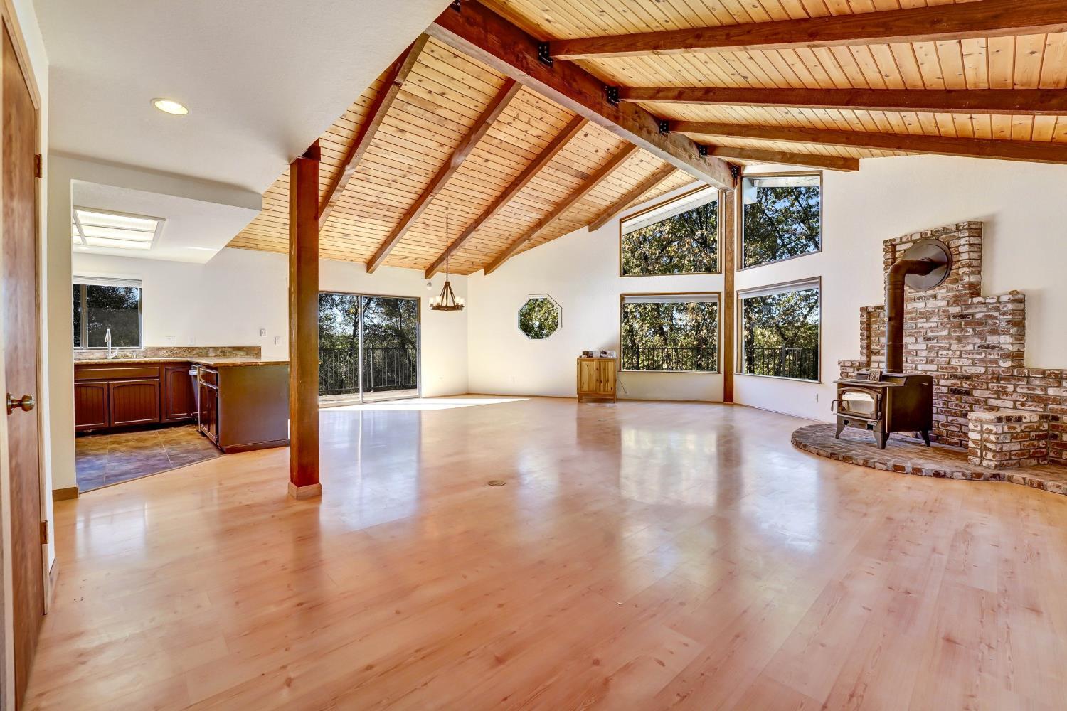 Living room with a vaulted wood ceiling, brick fireplace, and large windows overlooking trees.