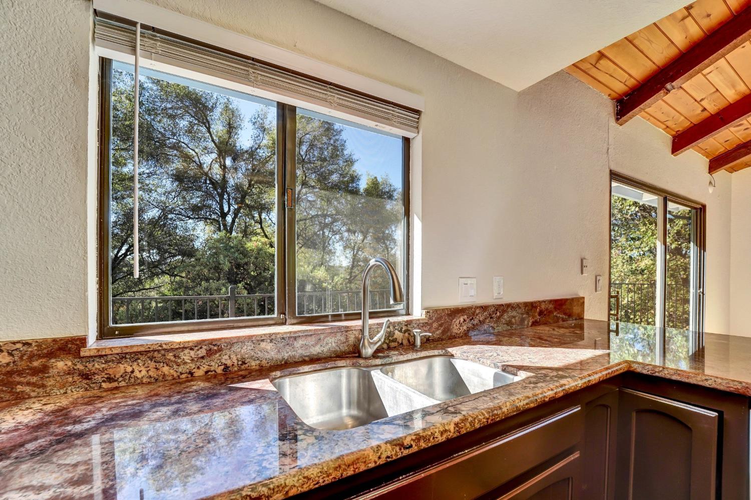 Kitchen sink and granite counters beneath large windows overlooking trees.