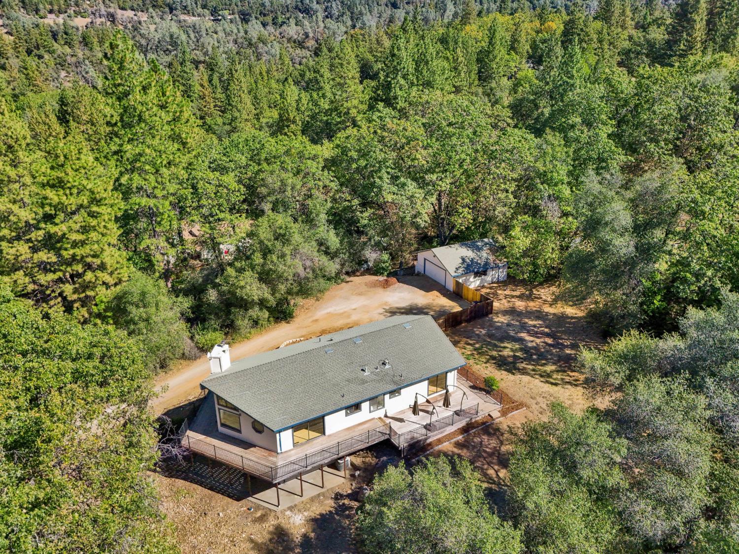 Aerial view of a home with a wraparound deck, detached outbuilding, and surrounding woodland.