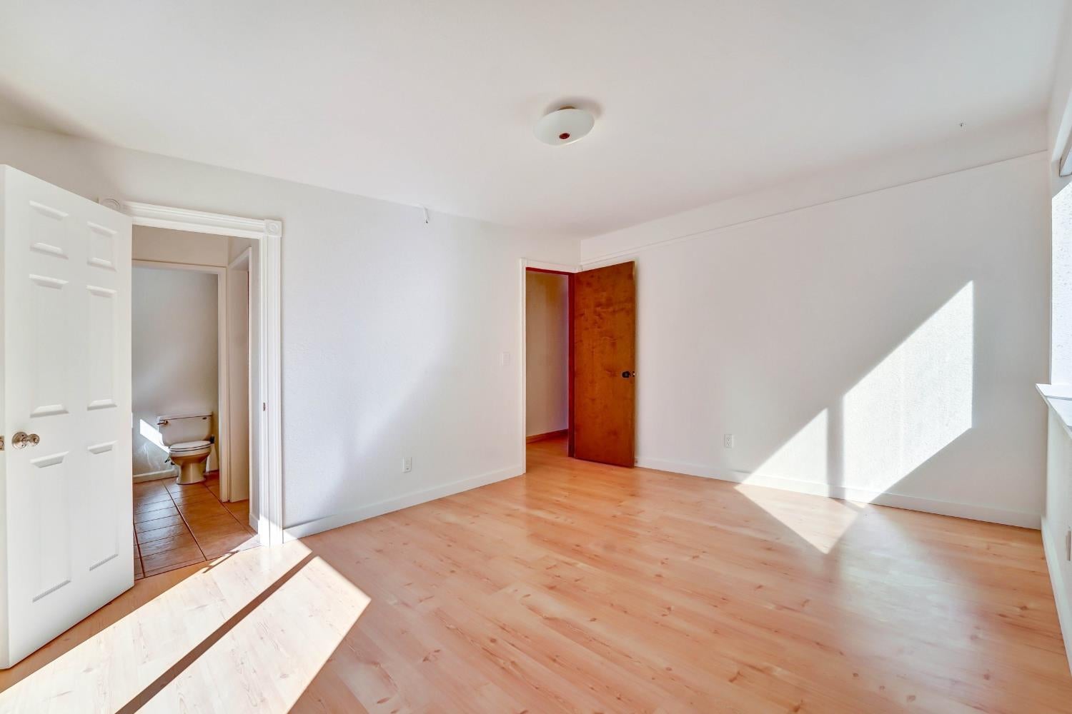 Empty room with light wood flooring, adjoining bathroom, and sunlight across the floor.