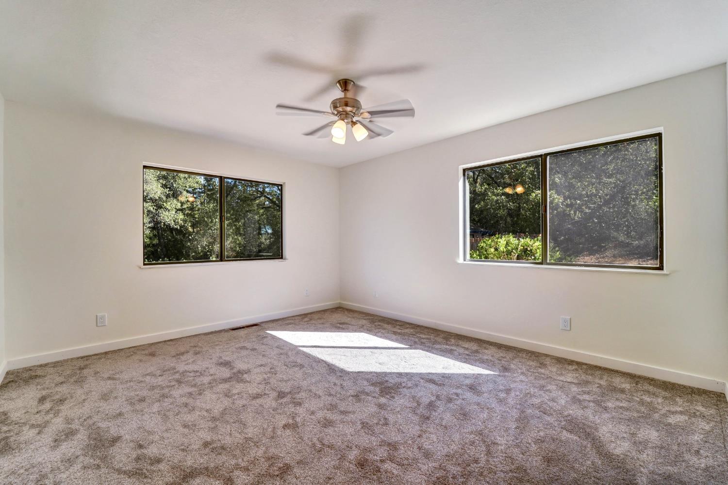 Unfurnished bedroom with taupe carpet, two large windows, and a ceiling fan with lights.