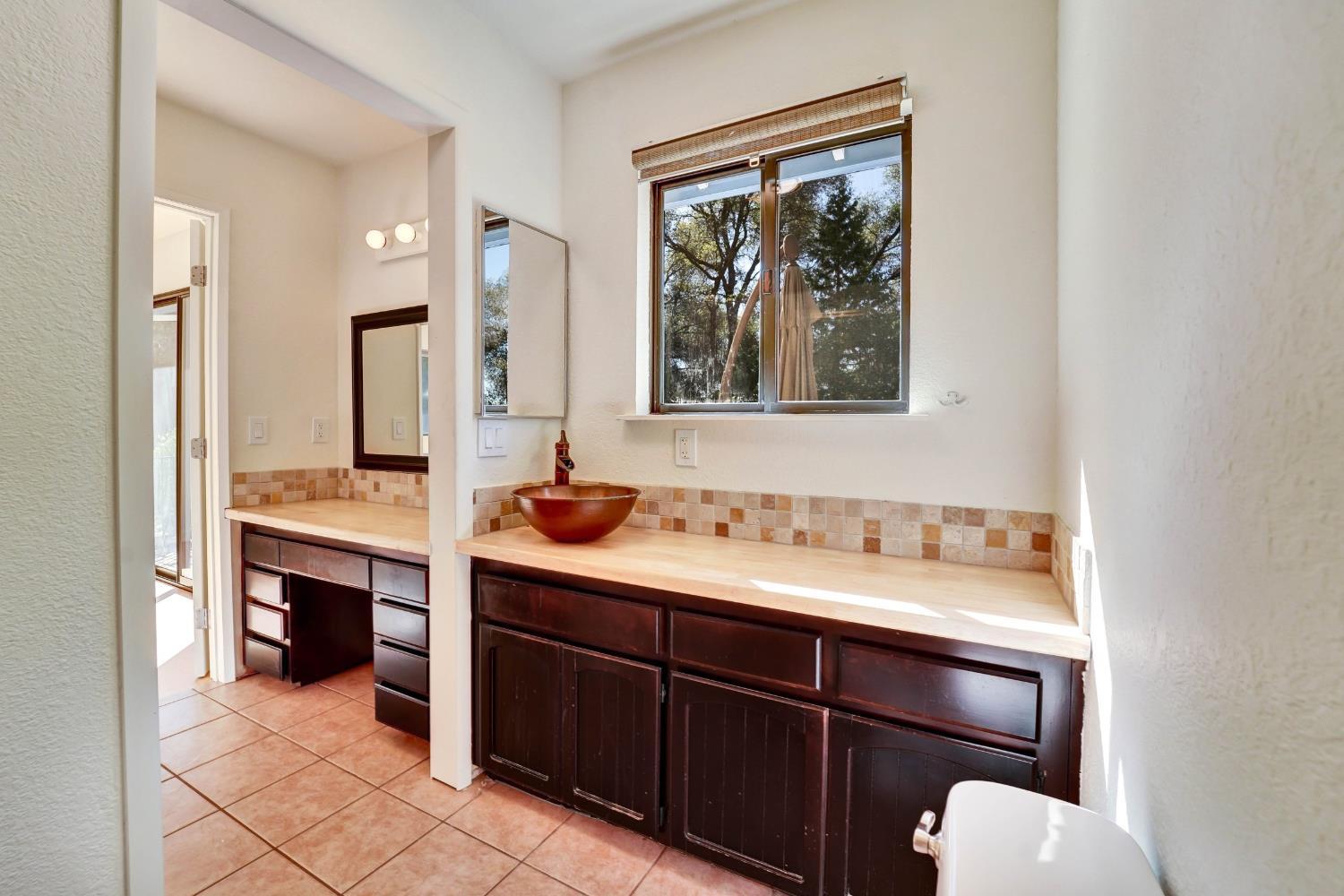 Bathroom vanity with a copper vessel sink, dark cabinets, tiled backsplash, and window overlooking trees.