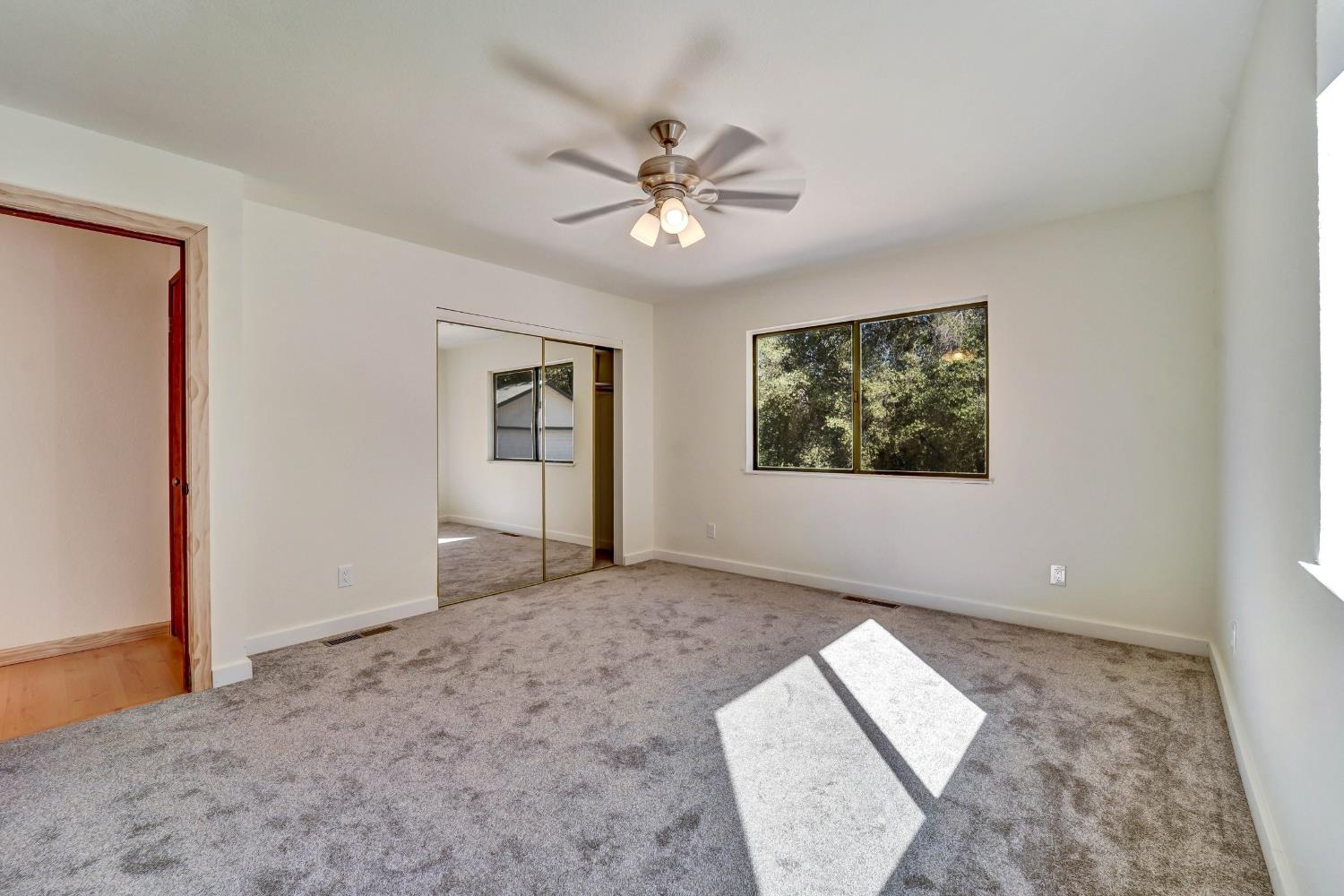 Empty carpeted bedroom with mirrored closet doors, a ceiling fan, and a window overlooking trees.