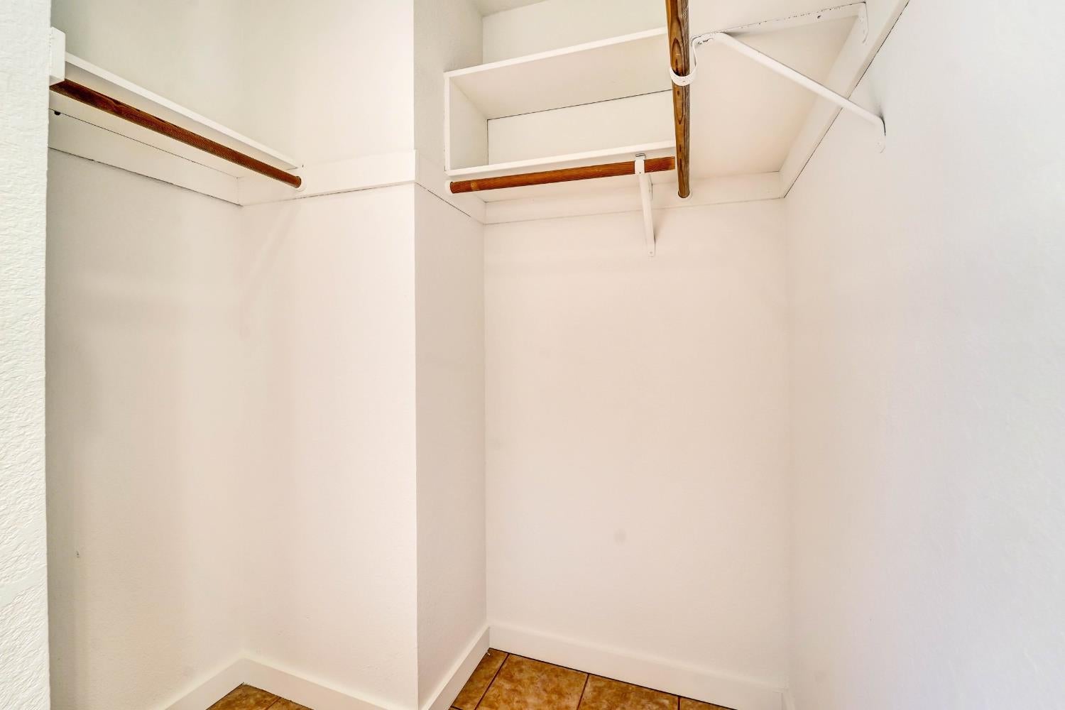 Empty walk-in closet with upper shelves, wooden hanging rods, and brown tile flooring.