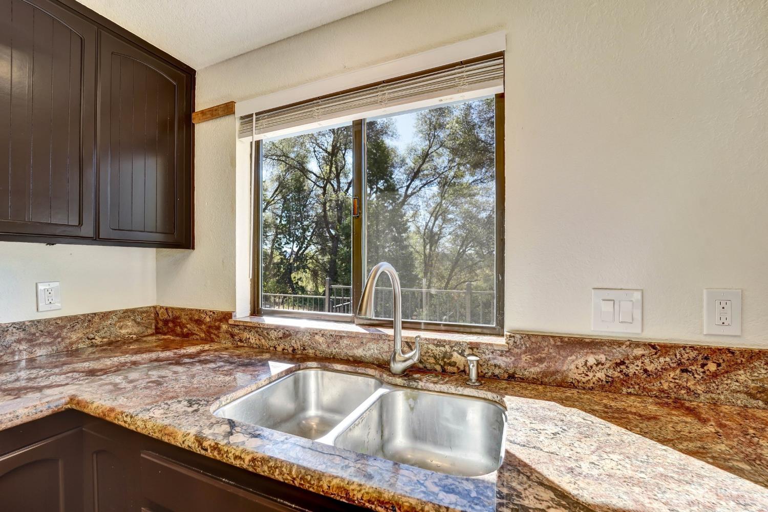Kitchen with dark wood cabinets, stone counters, and a double-basin sink beneath a window overlooking trees.