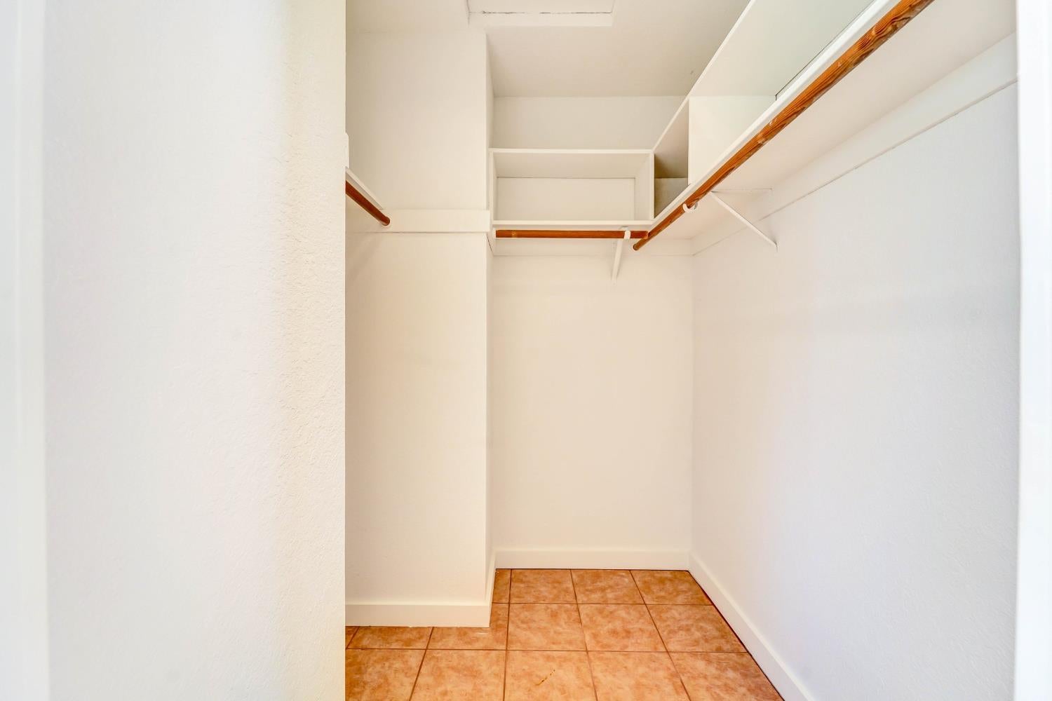 Walk-in closet with built-in shelving, wooden hanging rods, and terracotta-colored tile flooring.