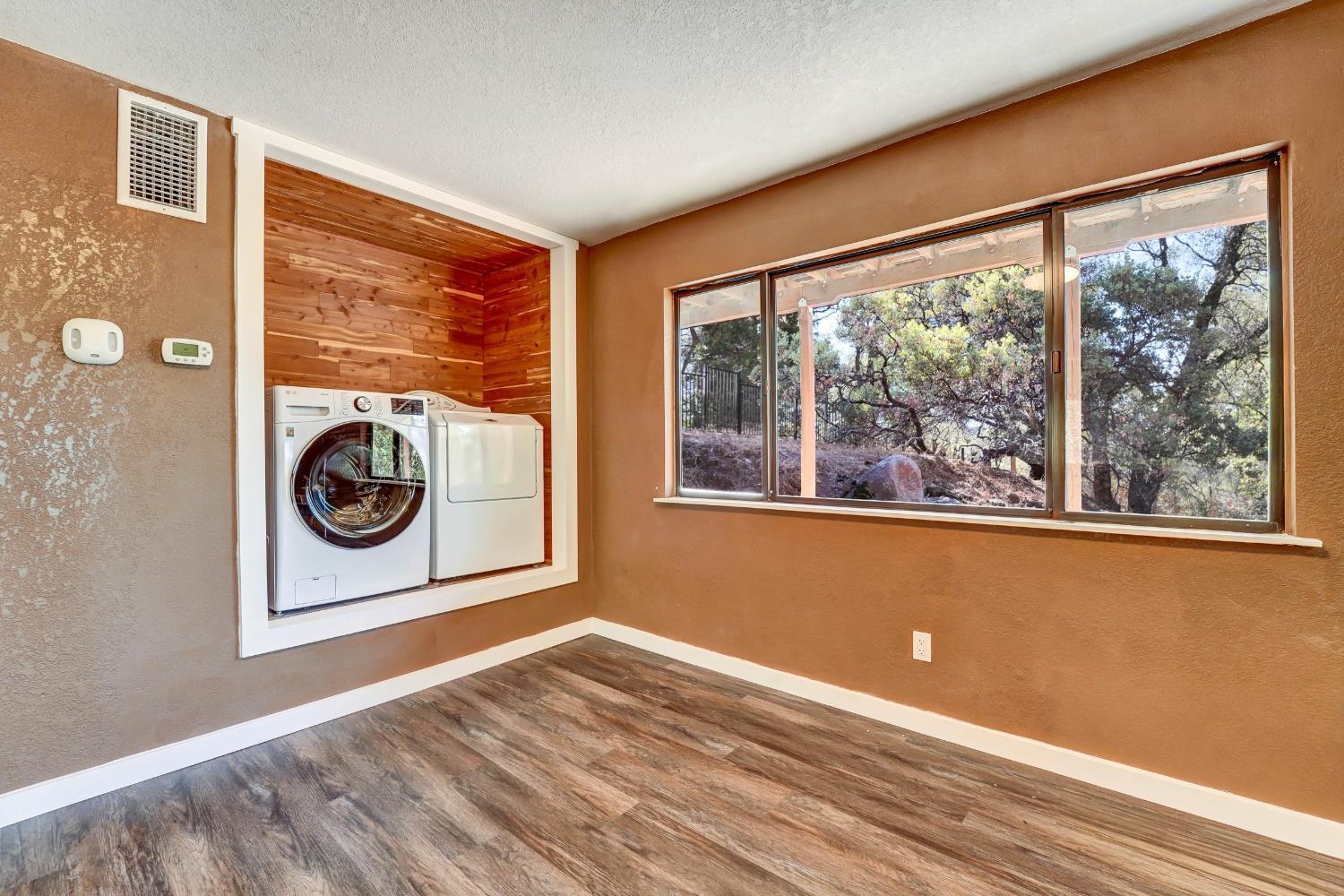 Laundry room with washer and dryer in a wood-paneled alcove beside large windows overlooking trees.