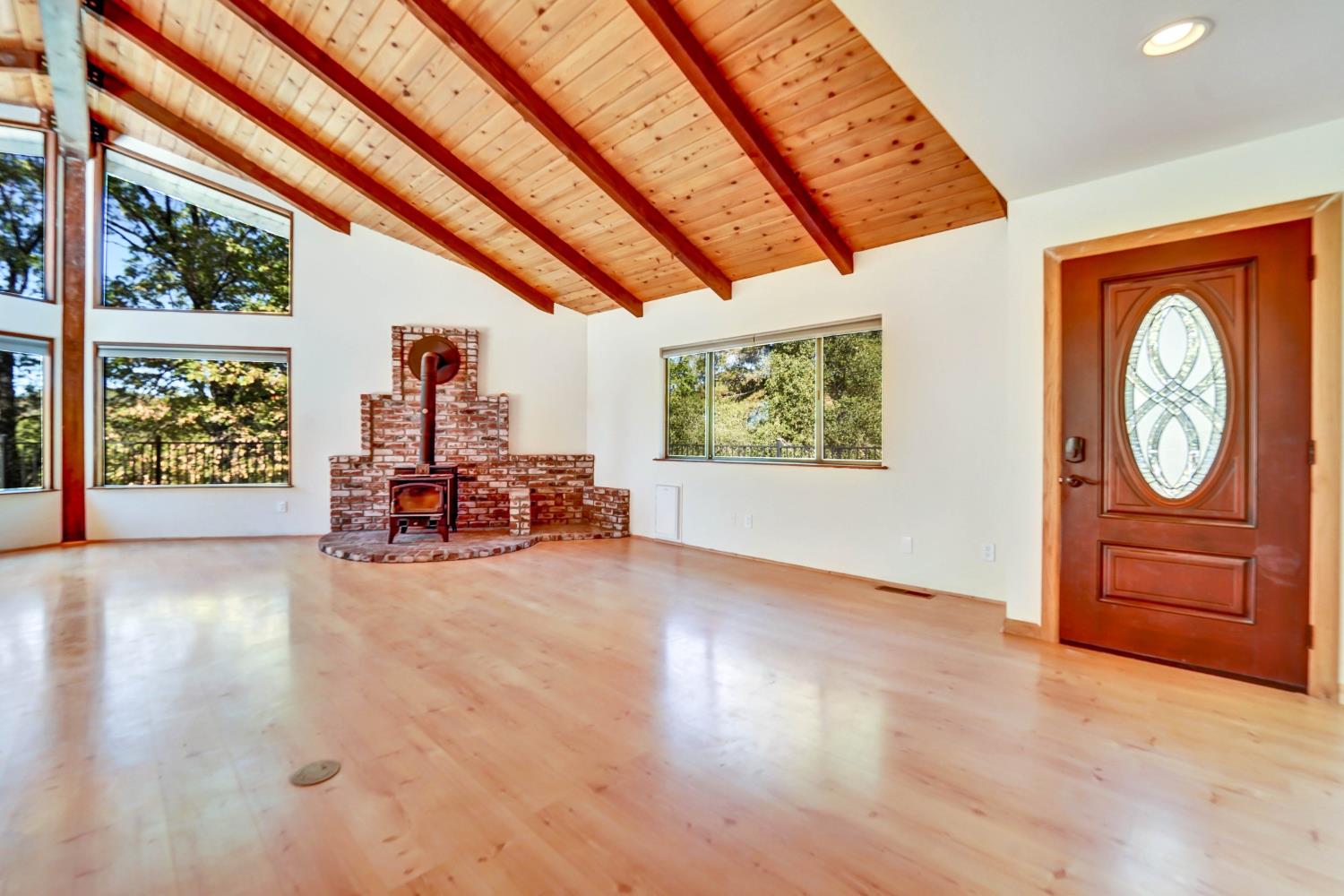 Vacant living room with a vaulted wood ceiling, brick fireplace and wood stove, and tall windows overlooking trees.