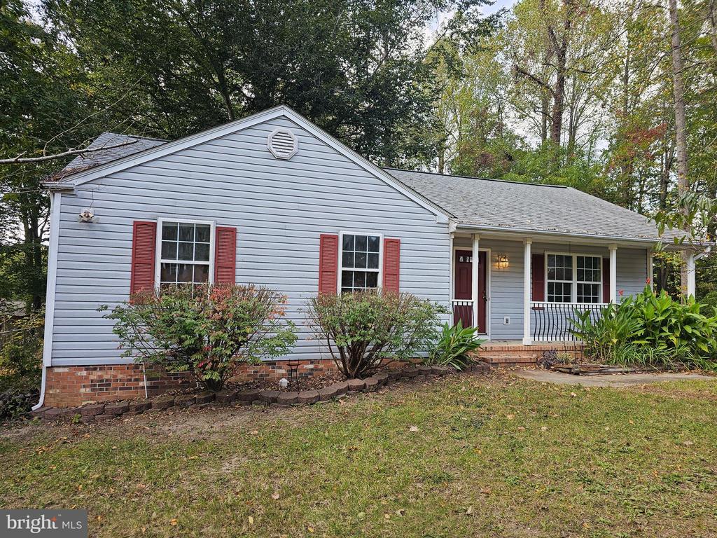 Single-story house with red shutters, a covered porch, and a brick foundation among trees.