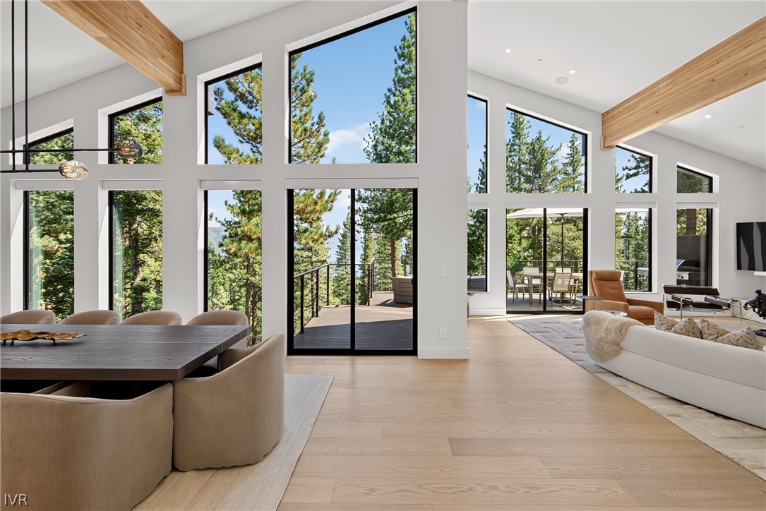 Open-plan living and dining room with tall windows overlooking pine trees and a deck.