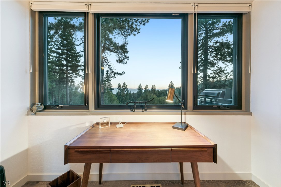 Wooden desk beneath three large windows overlooking pine trees and distant hills.