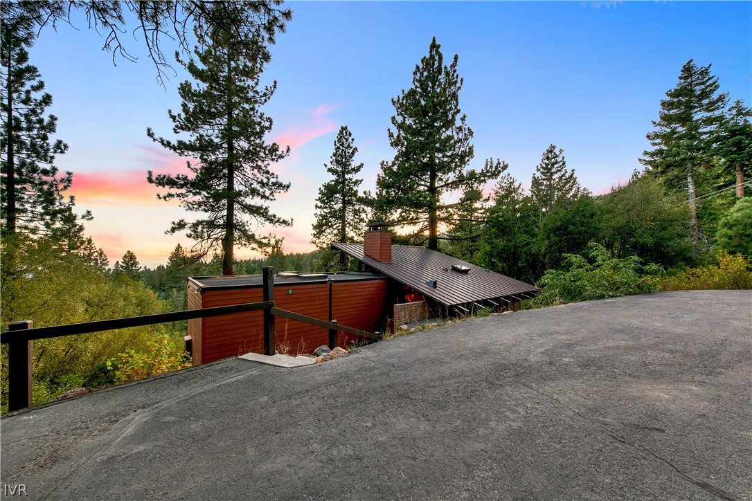 Red-sided home among tall pine trees beside a sloping paved driveway at sunset.