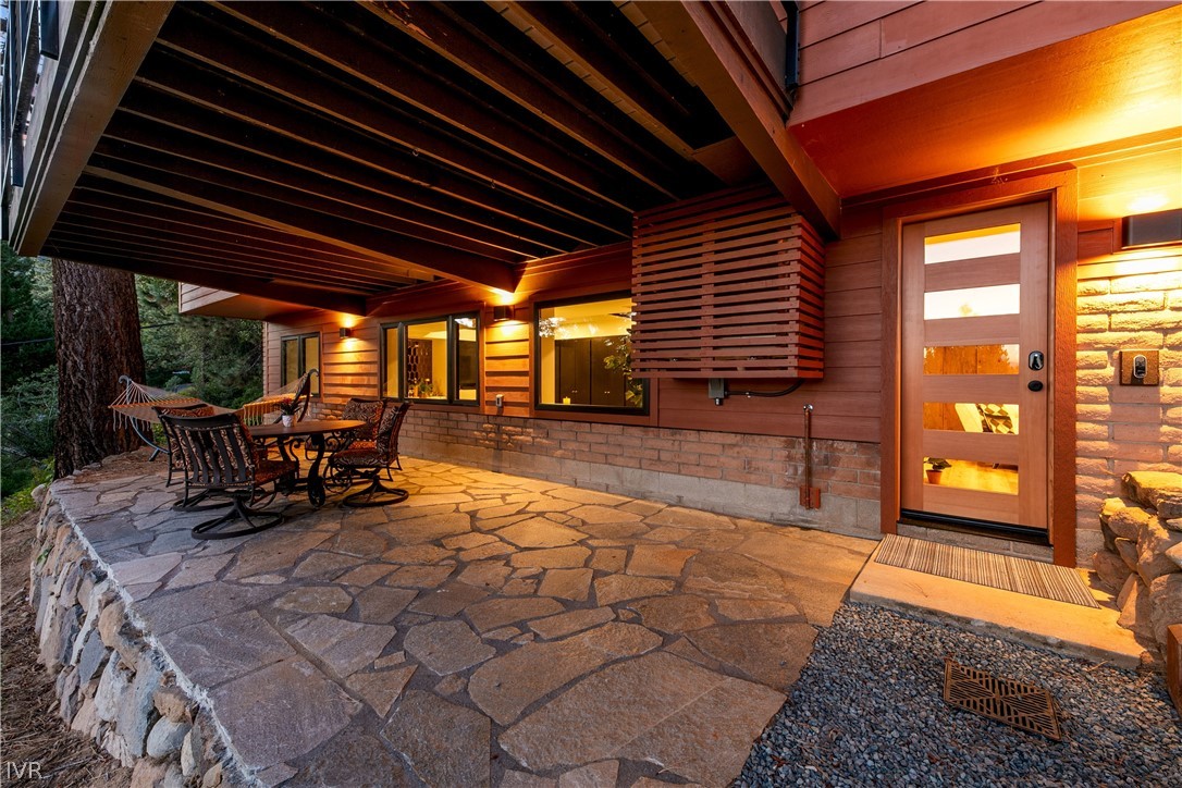 Covered stone patio with outdoor dining furniture and a warmly lit, glass-paneled entrance.