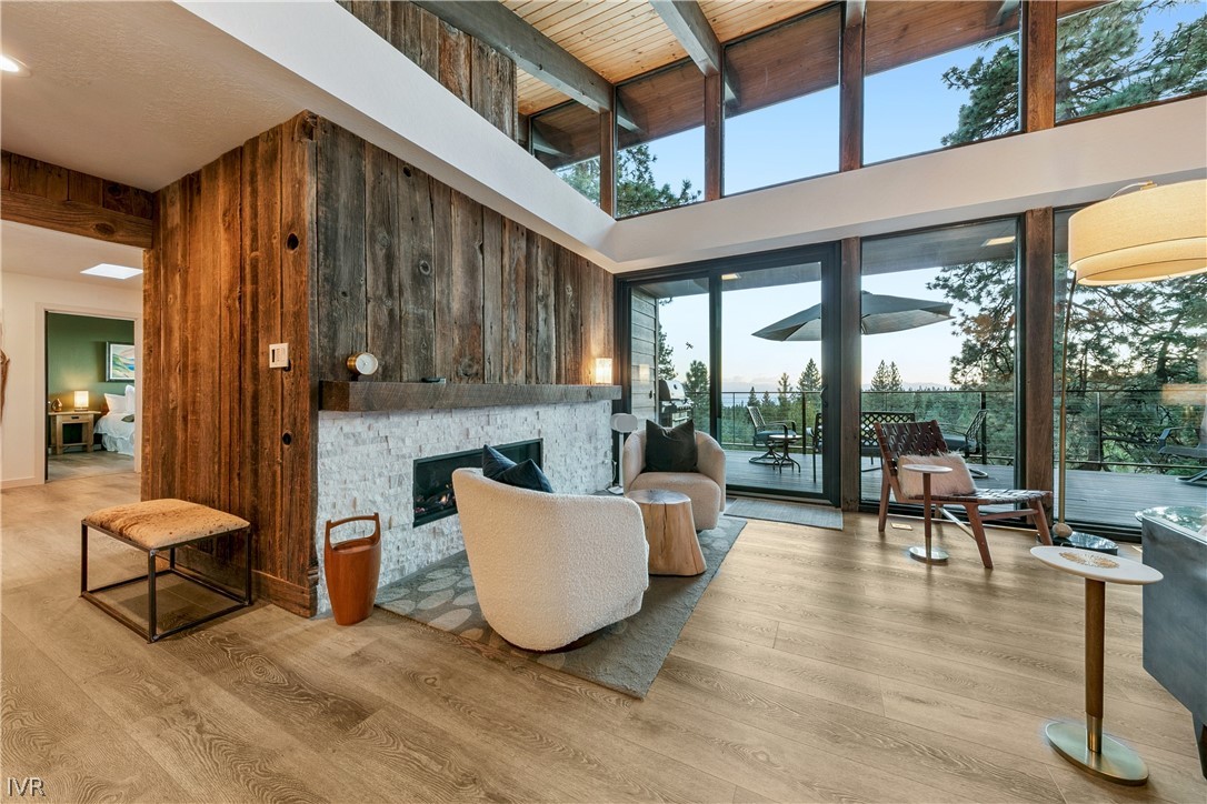 Living room with a stone fireplace, vaulted wood ceiling, and large windows overlooking trees.