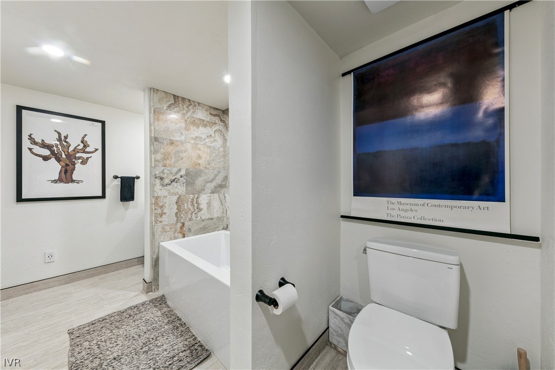 Bathroom with a white bathtub, stone-look tiled wall, and separate toilet beneath framed artwork.
