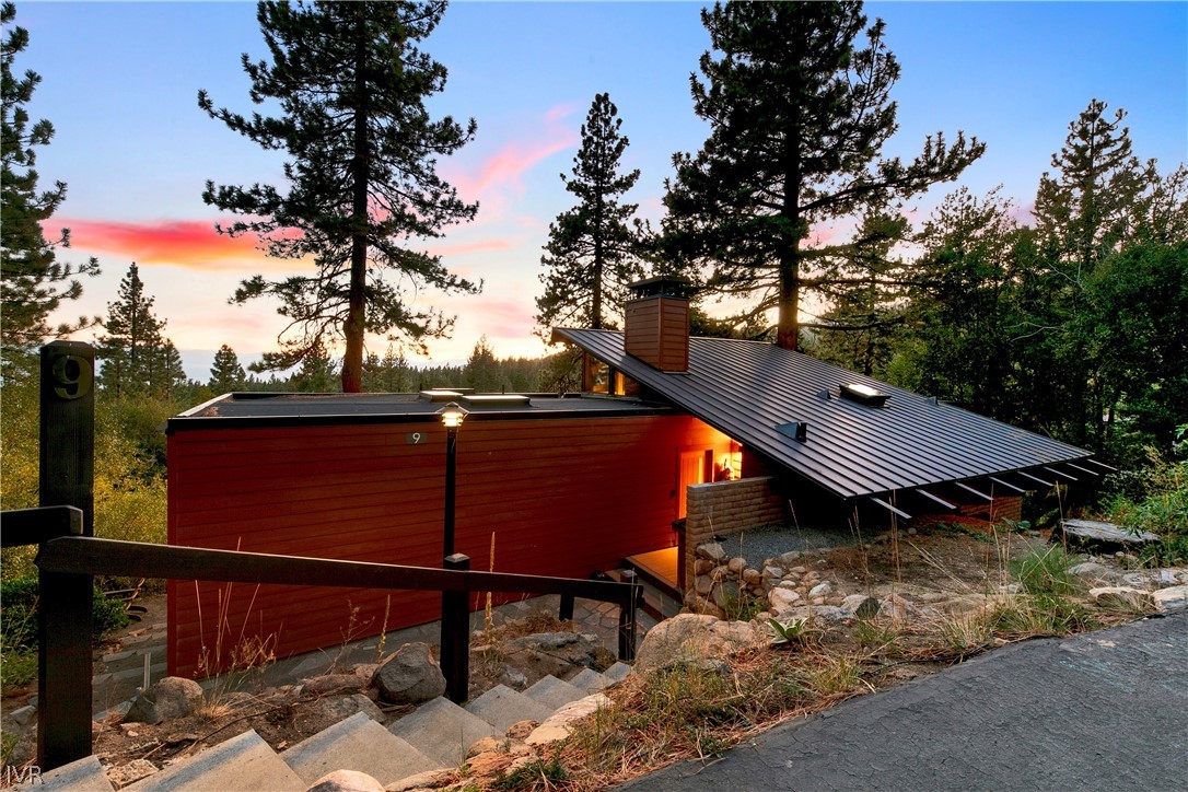 Modern red-sided home among pine trees, with a broad metal roof and stone steps at sunset.