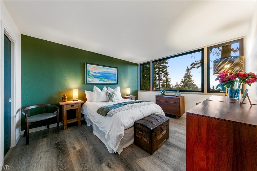 Staged bedroom with a white bed, deep green accent wall, and large windows overlooking pine trees.
