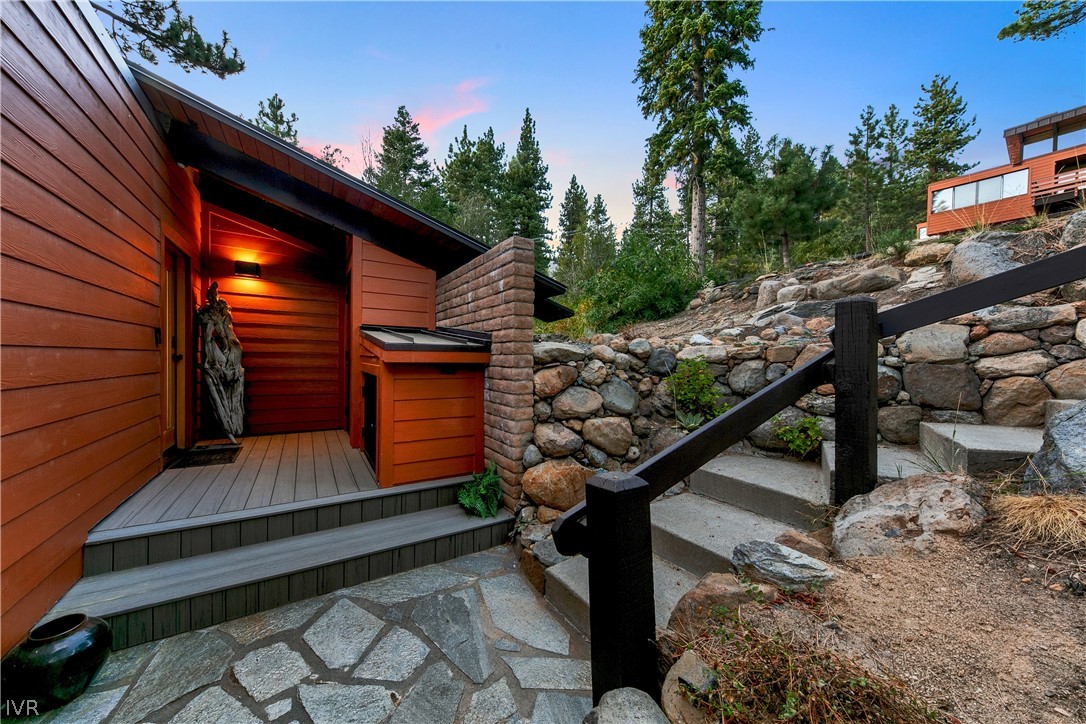 Wood-sided home entrance beside stone steps and a rocky, pine-covered slope.