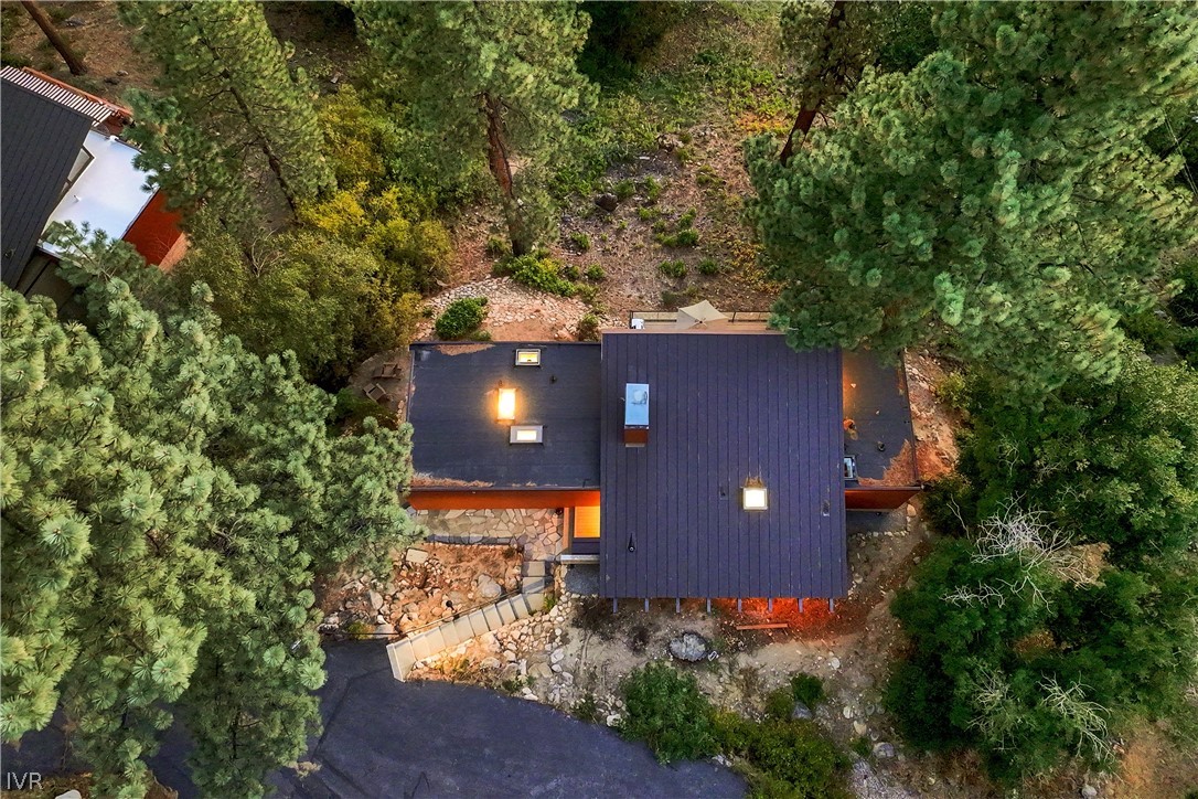 Aerial view of a dark-roofed home surrounded by pine trees, with stone paths and a rocky yard.