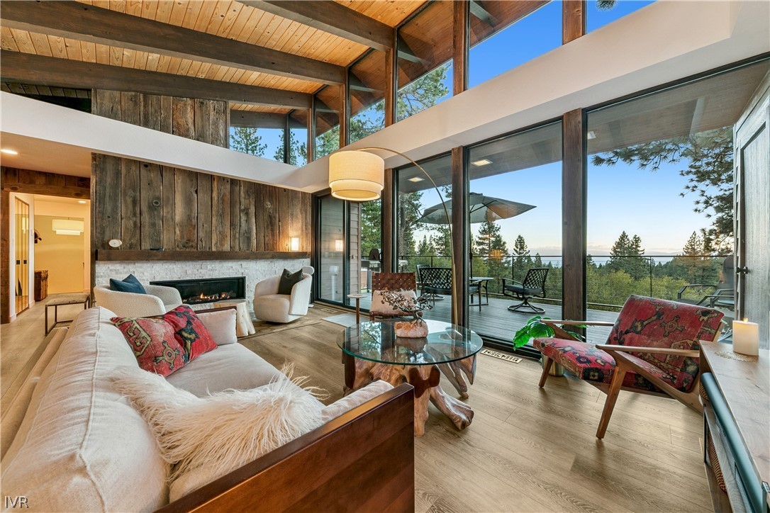 Living room with vaulted timber ceiling, stone fireplace, and deck windows overlooking trees and Lake Tahoe.