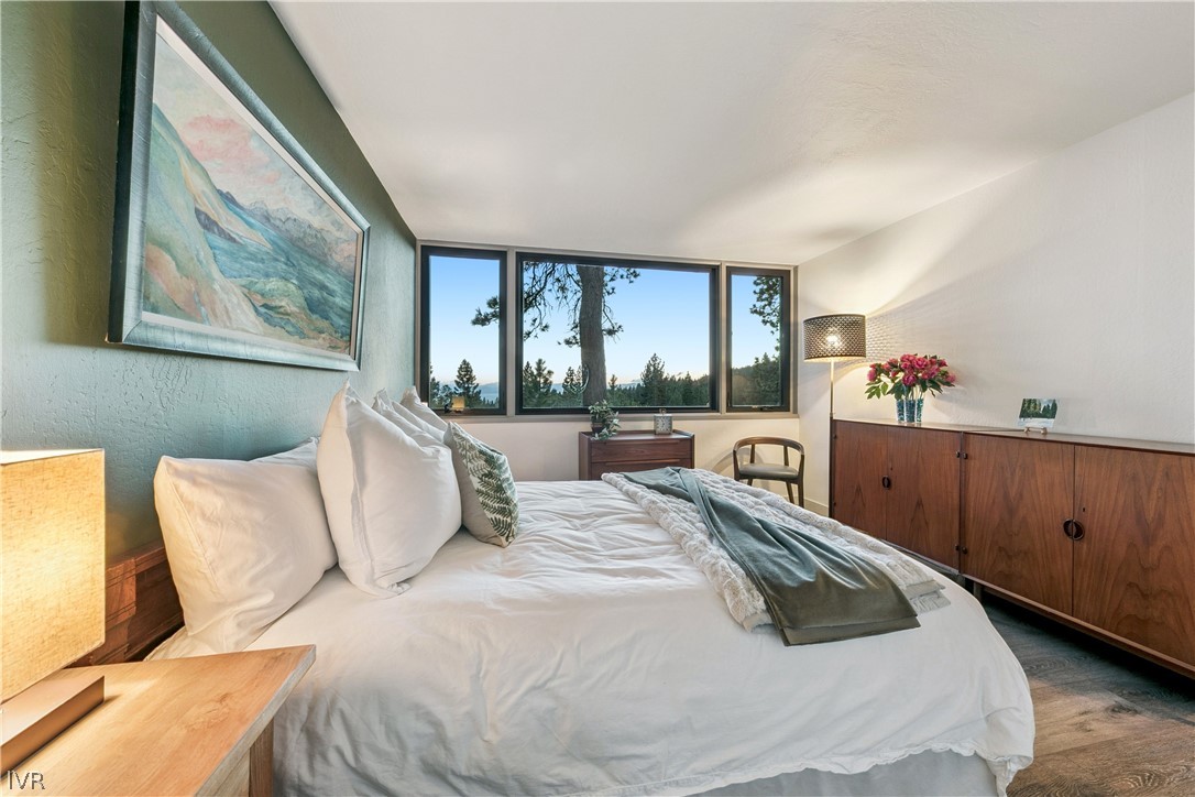 Bedroom with a white bed, wood cabinets, and wide windows overlooking trees and water.