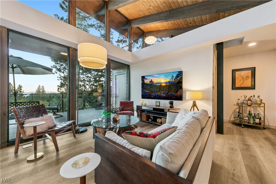 Living room with vaulted wood ceiling, tall windows, and a furnished deck among the trees.