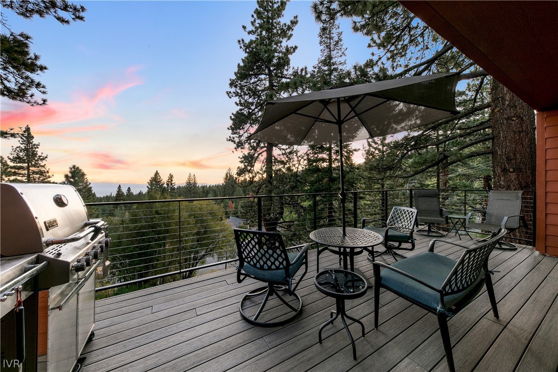 Furnished deck with a grill and umbrella overlooking pine trees at sunset.