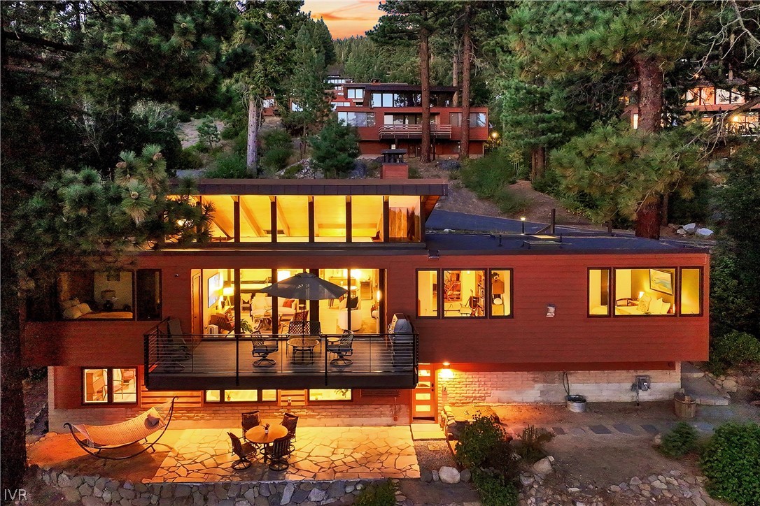 Modern wood-and-glass home glowing at dusk among pines, with a balcony deck and stone patio.