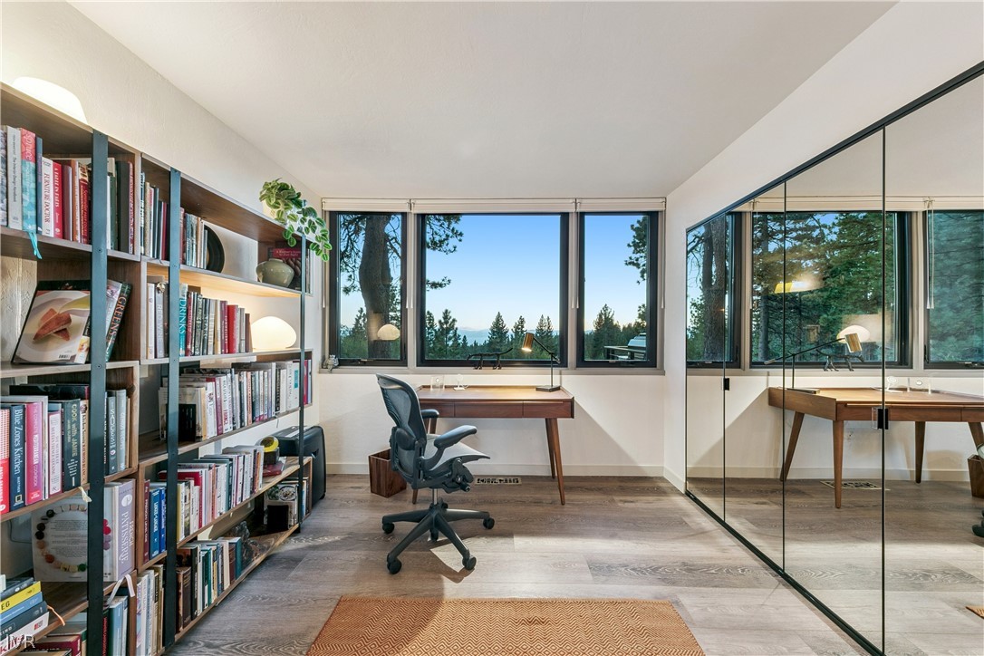 Furnished home office with wall-length bookshelves, a wooden desk, mirrored doors, and forest views through large windows.