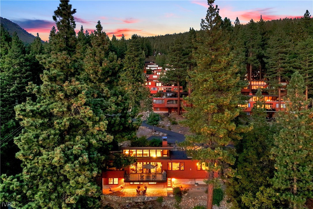 Aerial view of a warmly lit home among pine trees, with hillside homes and a pink dusk sky.