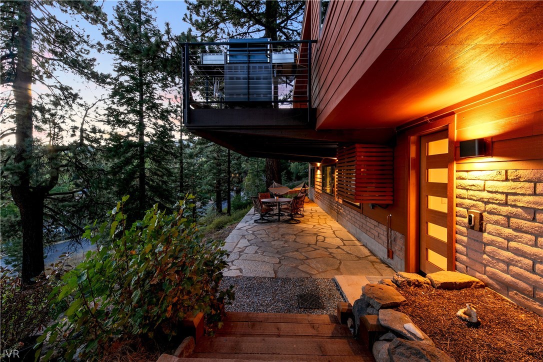 Modern wood-and-stone home with a warmly lit stone patio surrounded by pine trees at dusk.