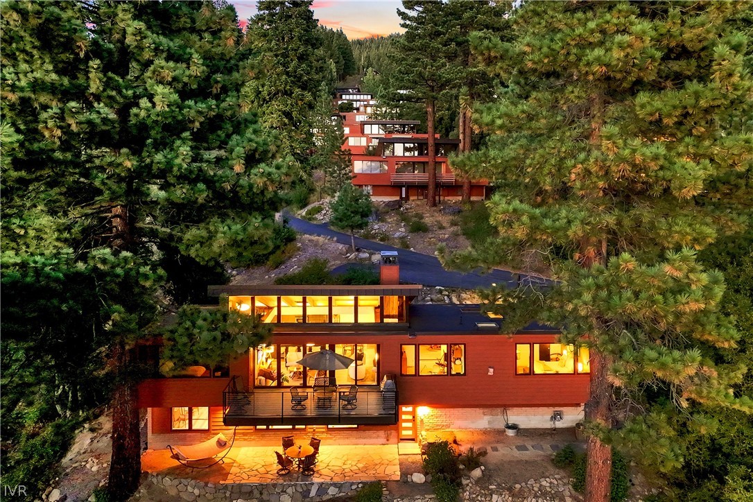 Redwood-sided home among pine trees, with glowing windows, a raised deck, and a stone patio.