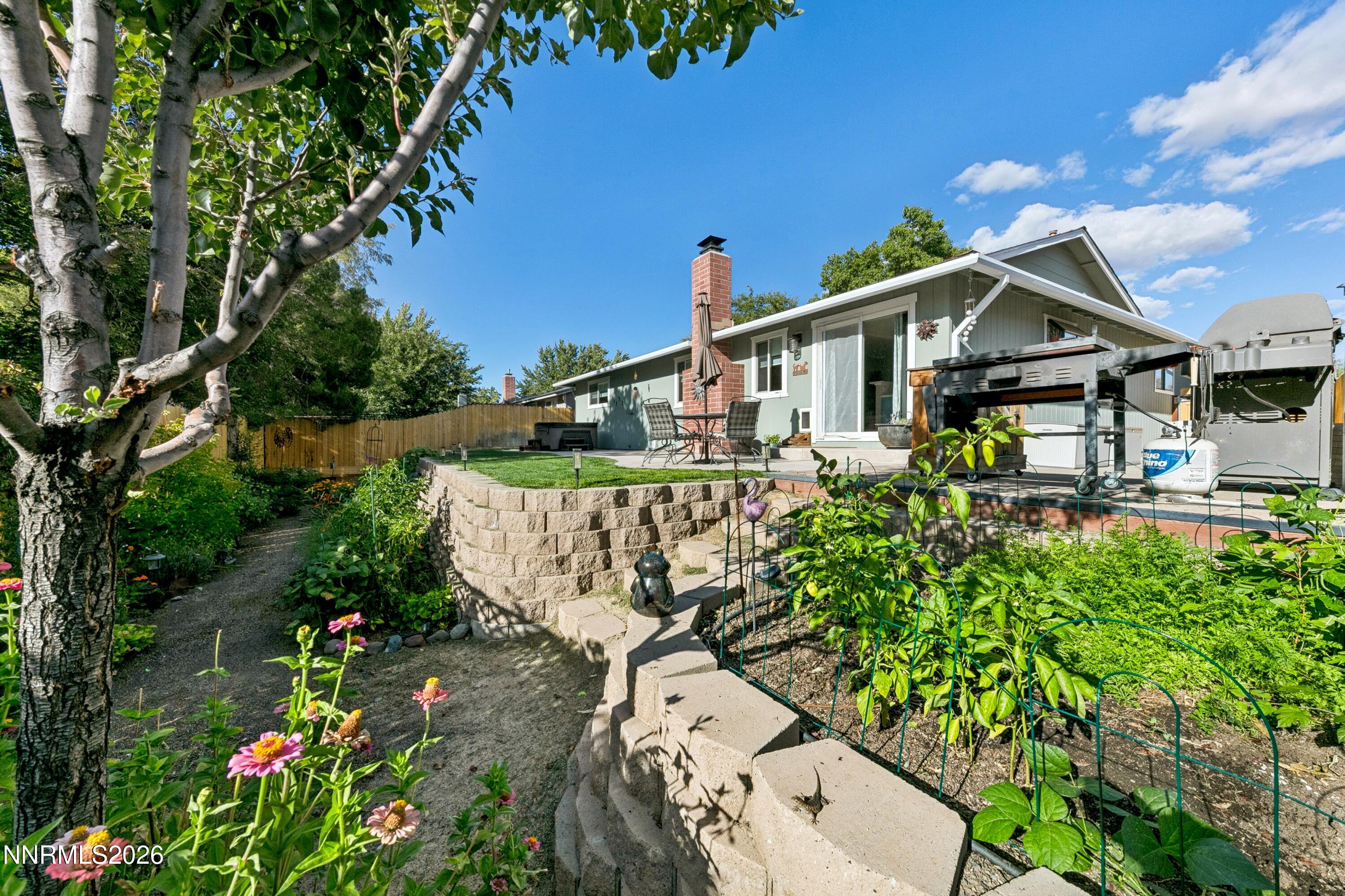 Backyard patio with brick chimney, barbecue grills, tiered garden beds, flowers, and mature trees.