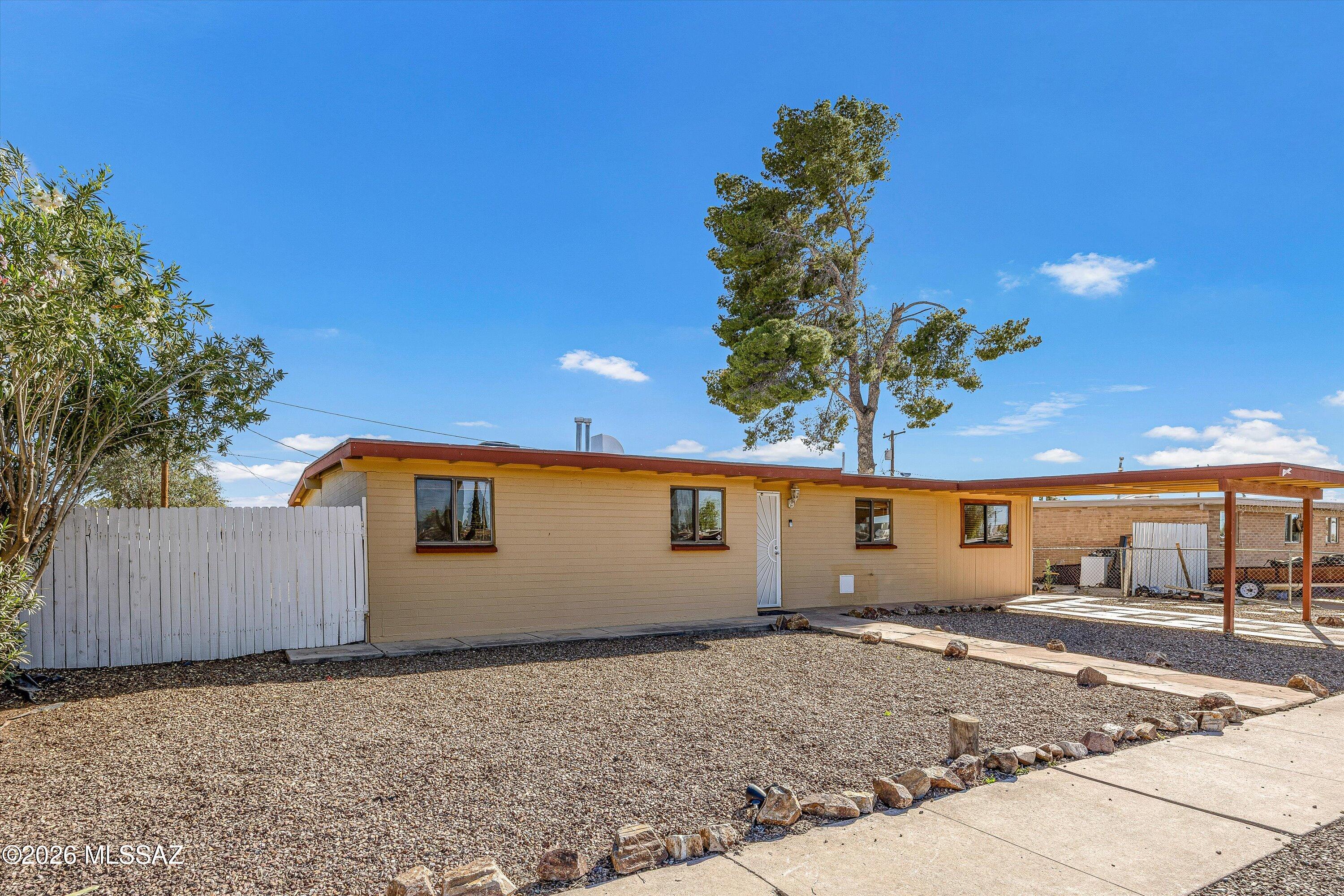 Single-story tan home with a covered carport, white fence, and gravel yard.