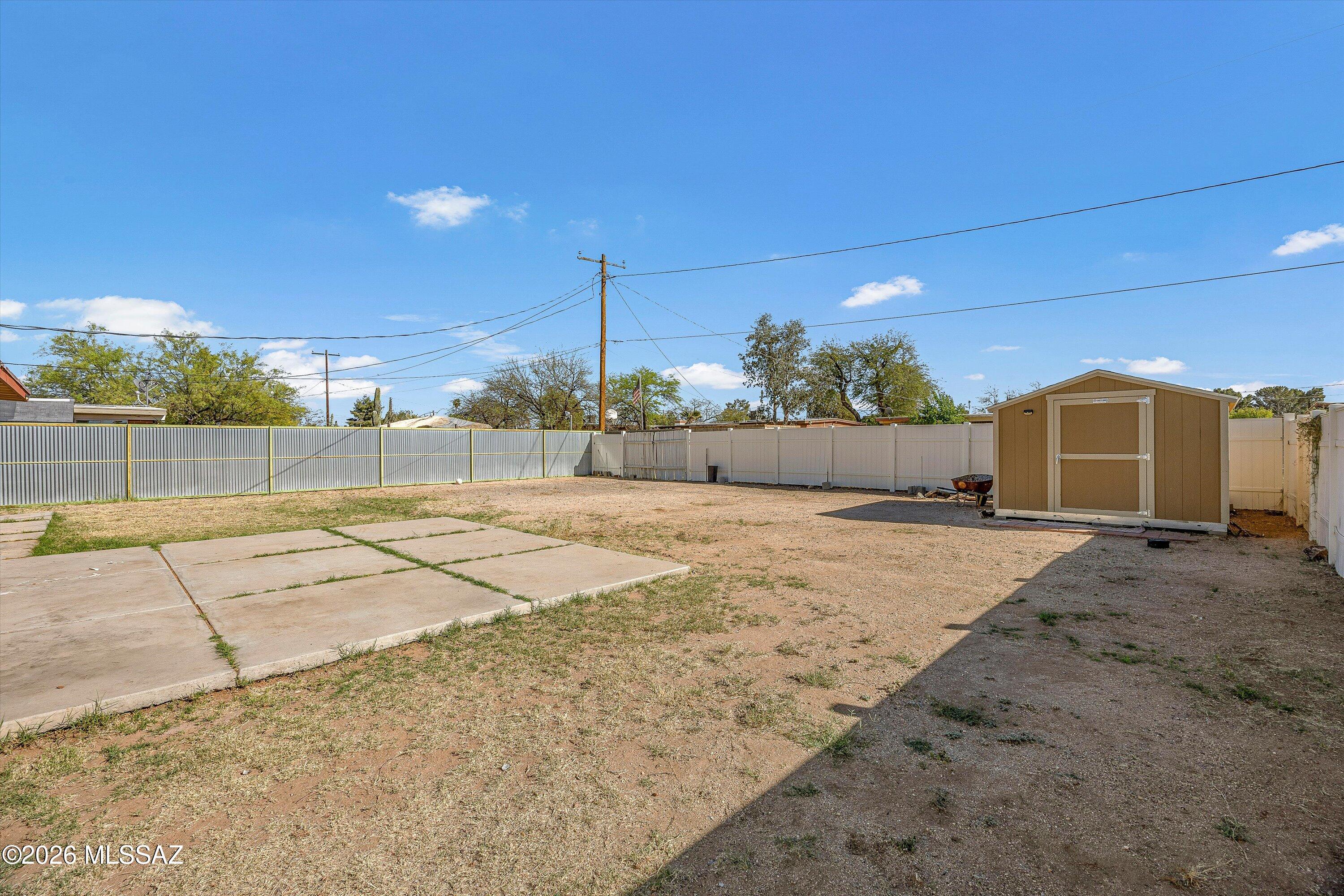 Fenced backyard with a concrete patio, sparse grass, and a storage shed.