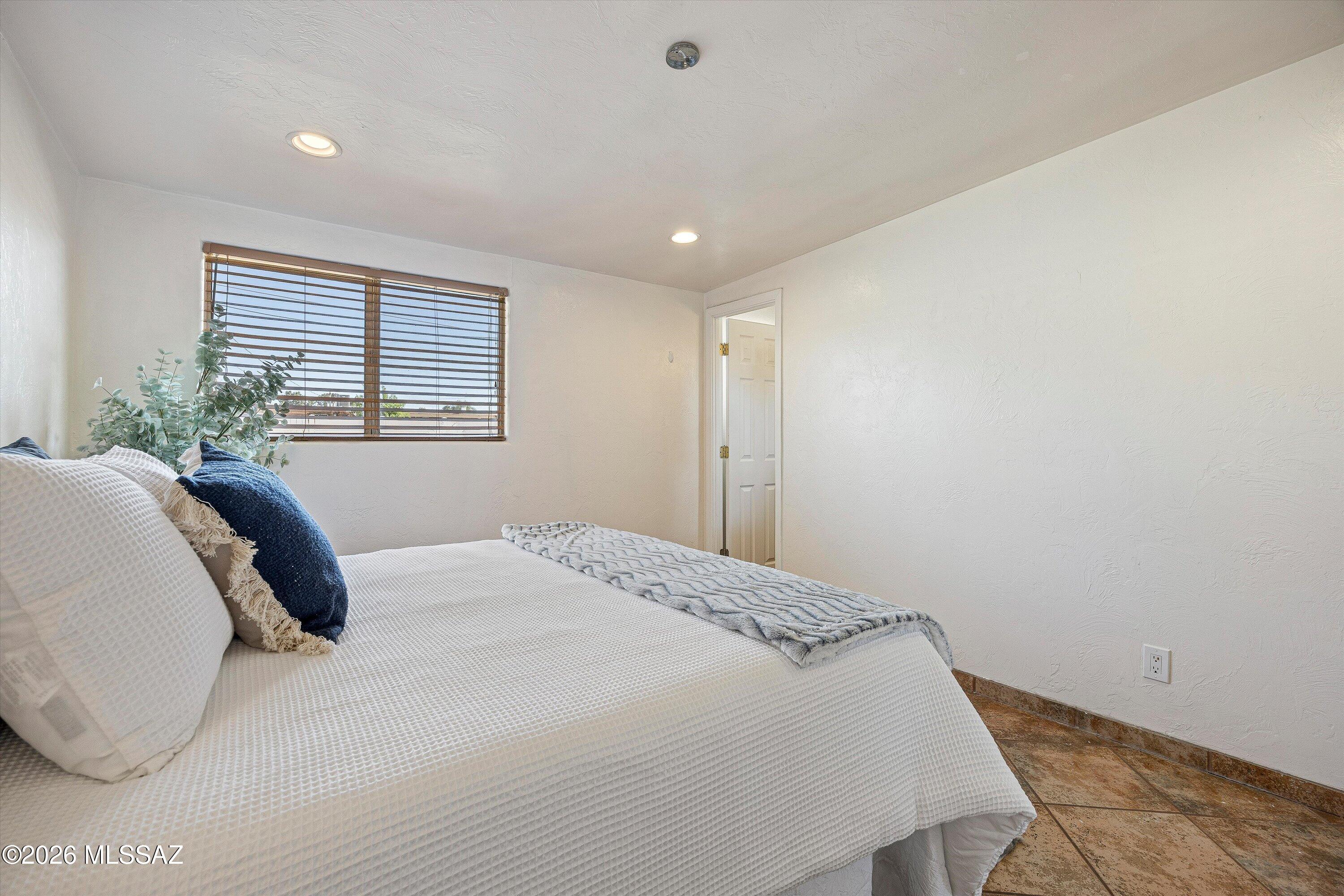 Bedroom with a white-covered bed, wooden window blinds, and brown tile flooring.