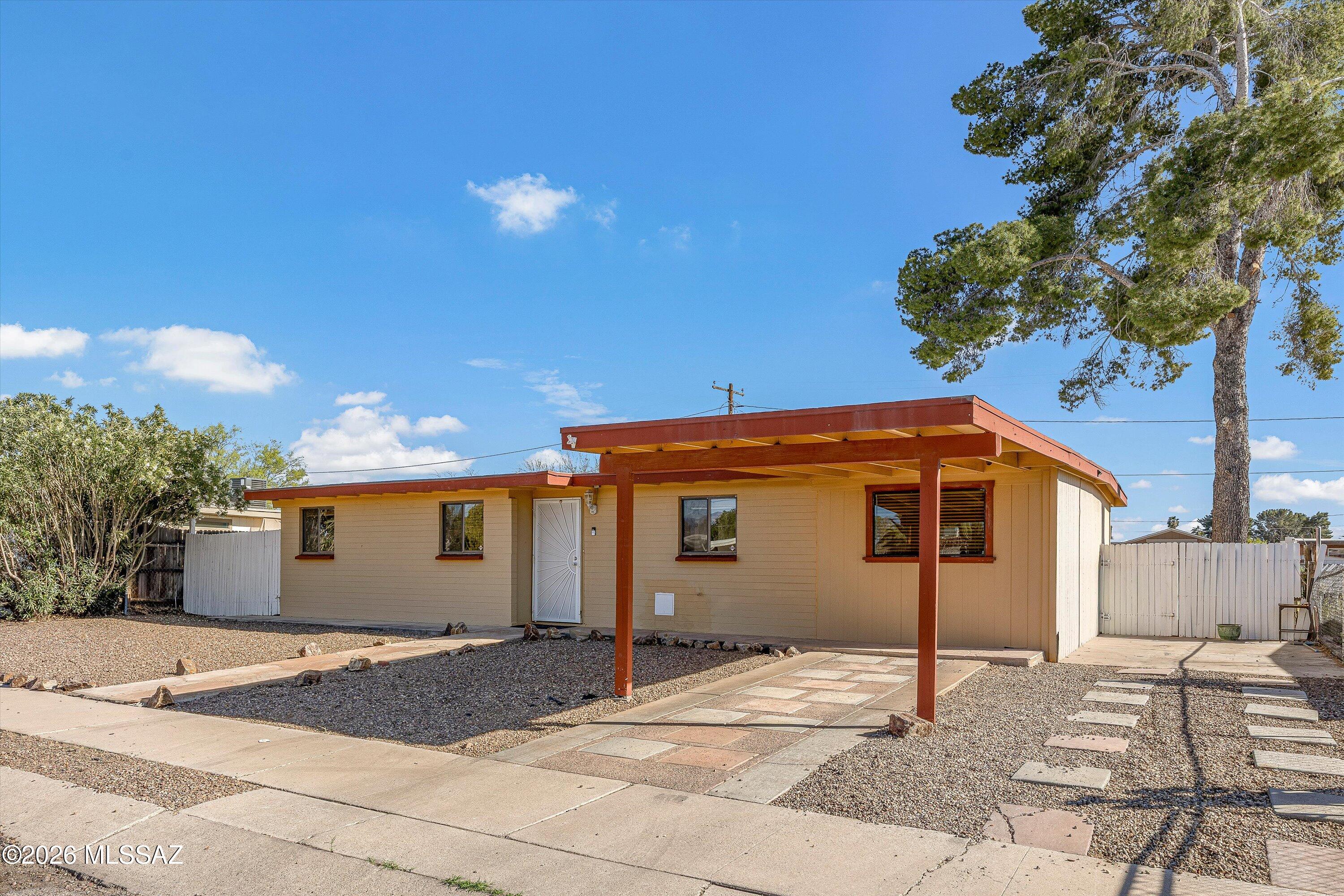 Single-story tan home with a broad covered carport, gravel yard, and white privacy fencing.