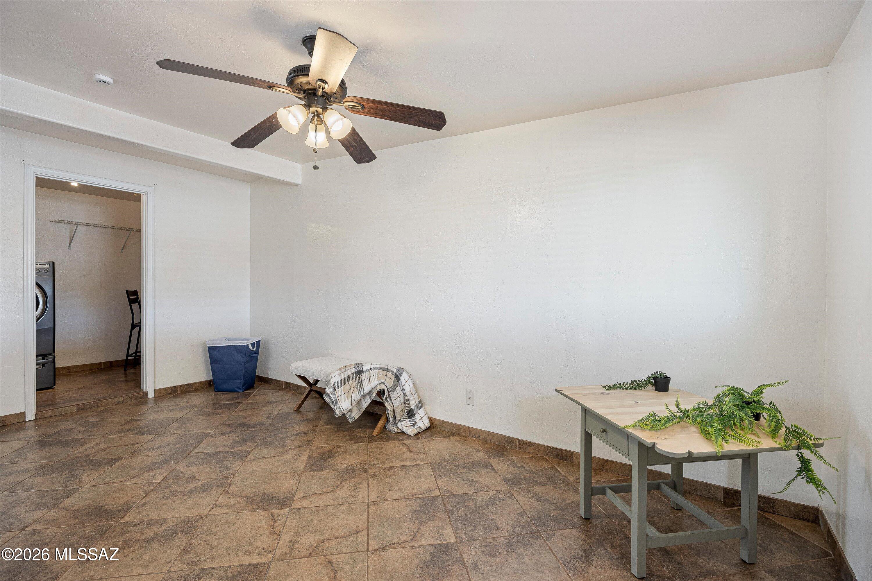 Tiled room with a ceiling fan, small bench, plant-covered table, and doorway to the laundry area.