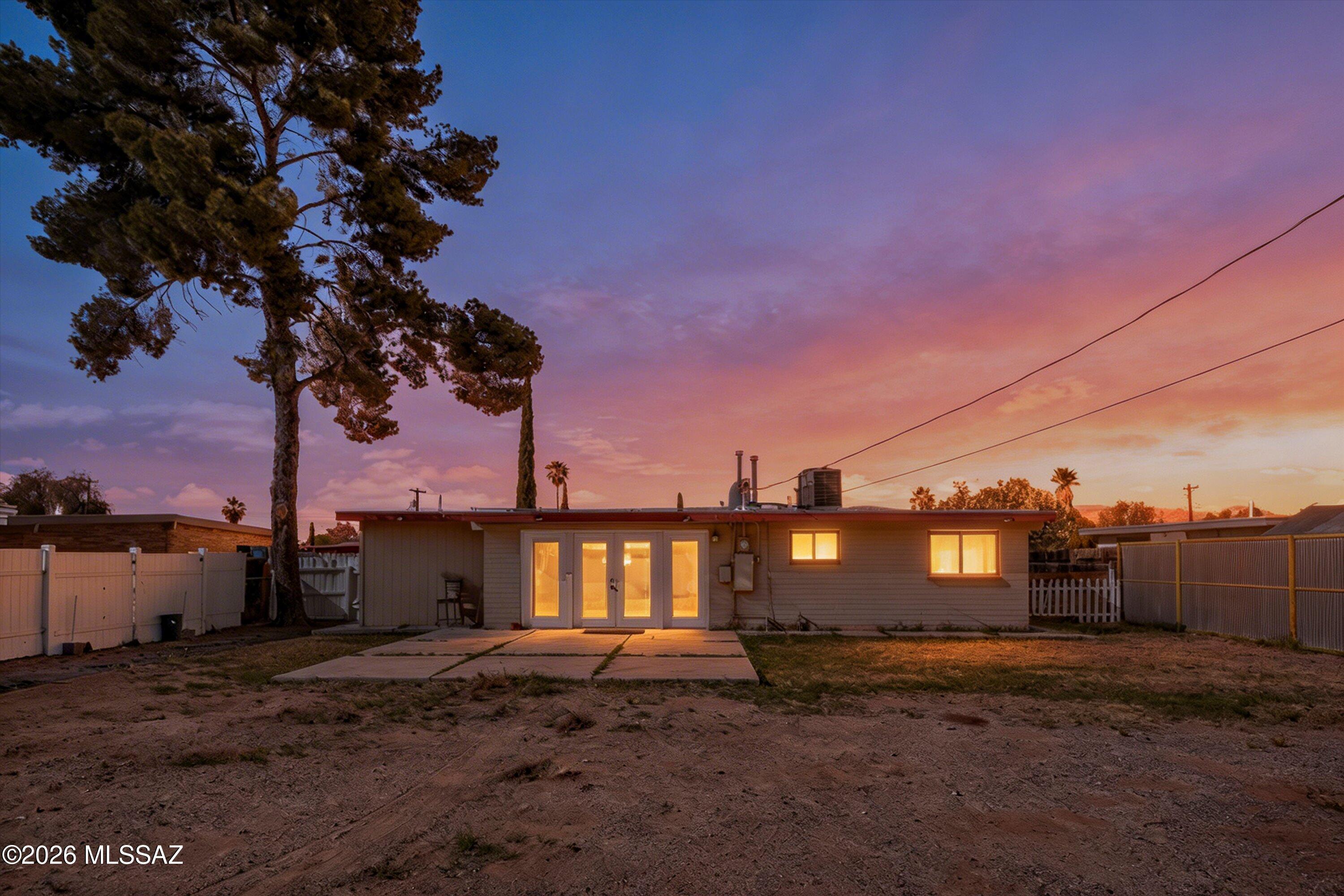 Single-story home's rear exterior at dusk, with glowing windows, a concrete patio, and a large tree.