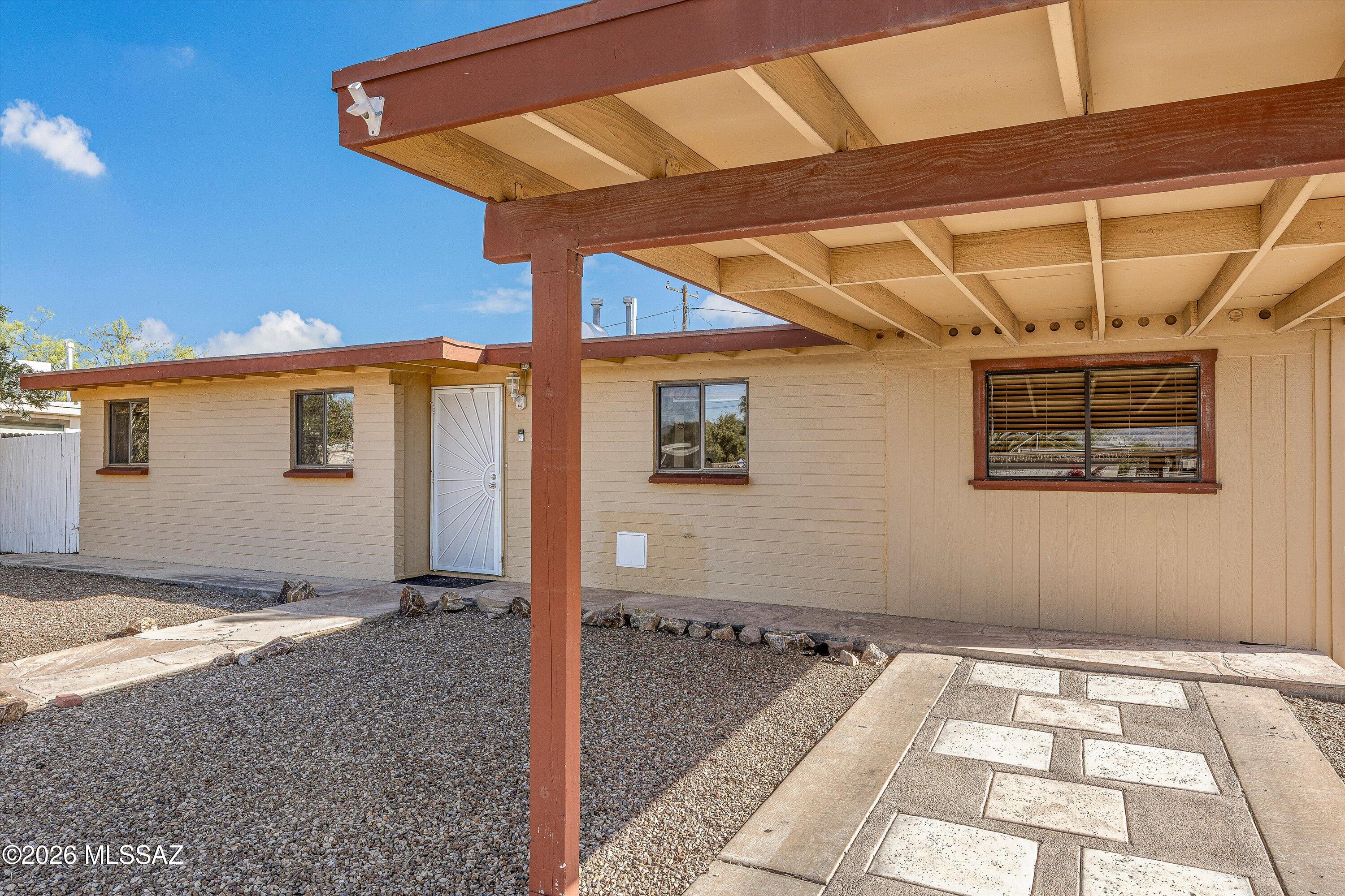 Single-story beige home with a covered porch, white entry door, gravel yard, and paved walkway.