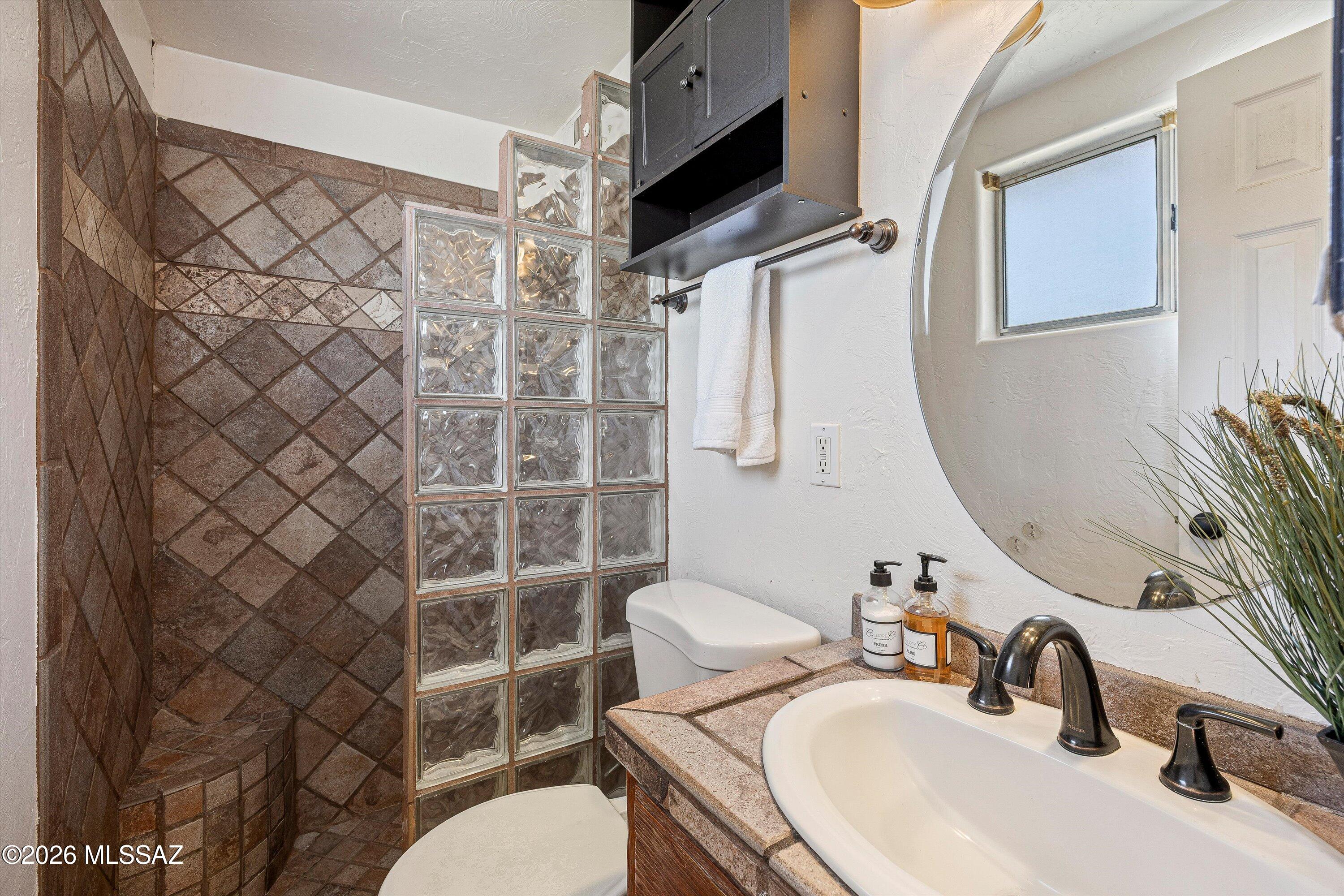 Bathroom with a brown tile shower, glass-block divider, and stone vanity with round mirror.