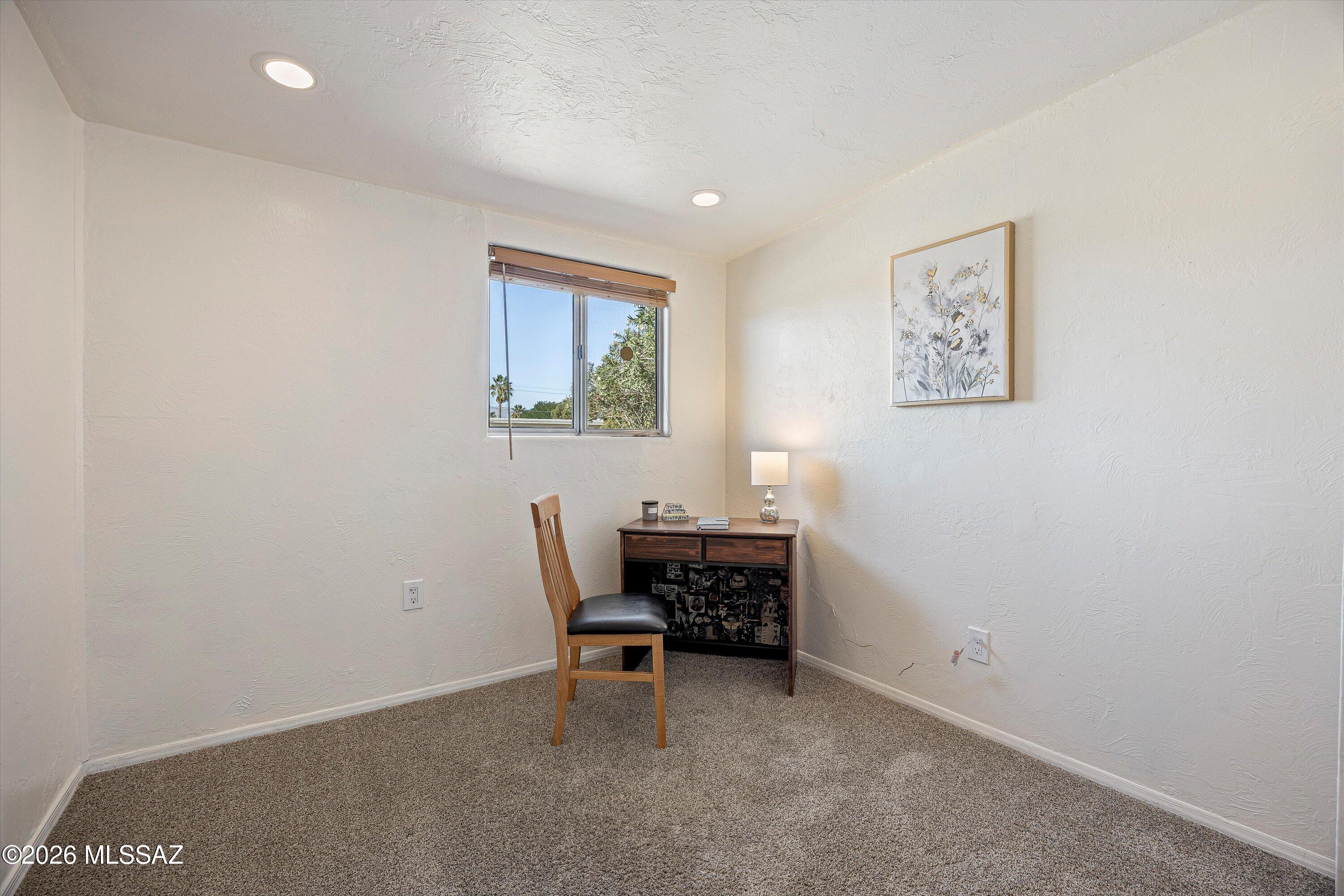 Carpeted room furnished with a wooden desk and chair beside a window with wooden blinds.