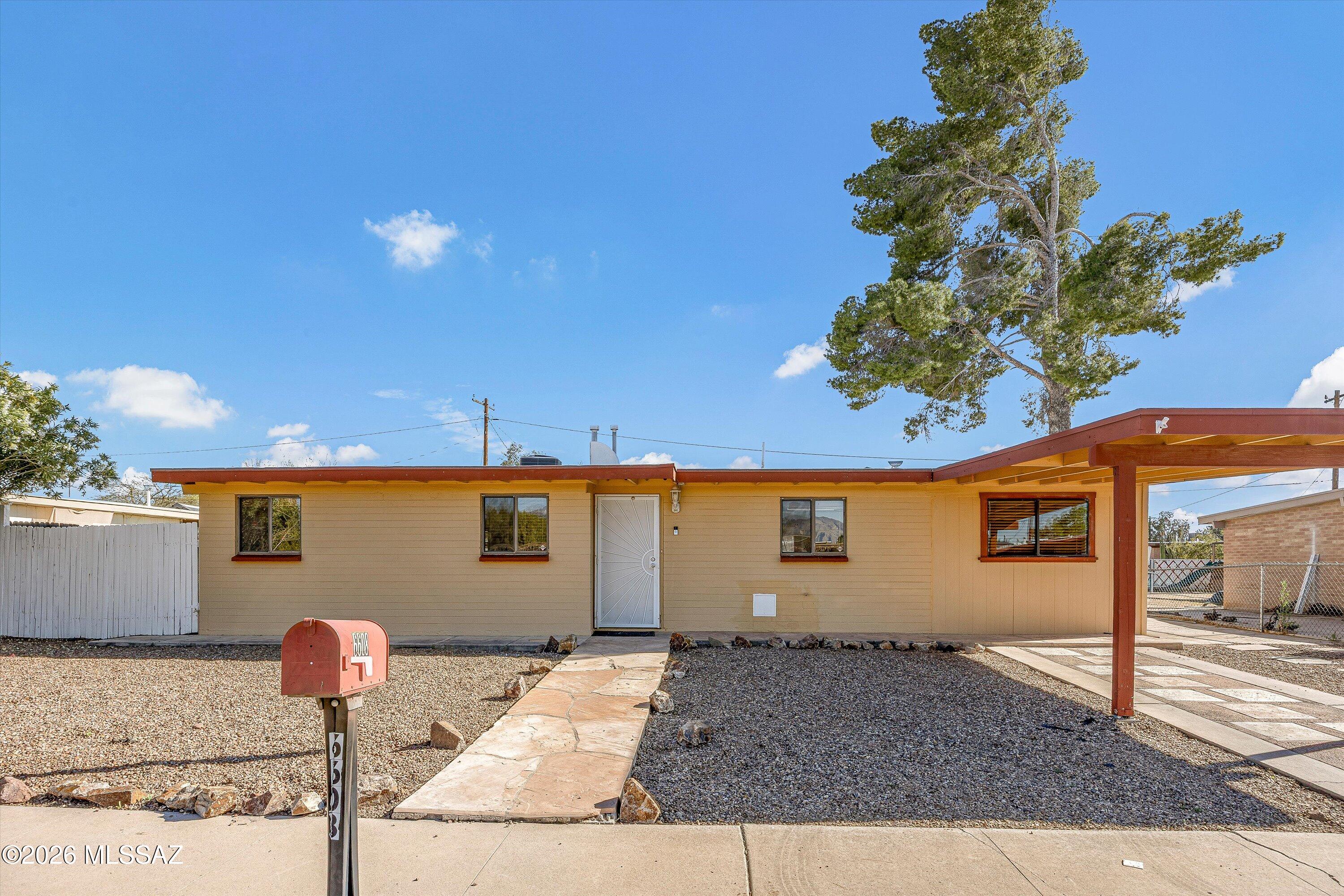 Single-story tan home with red-brown trim, gravel landscaping, and an attached covered carport.