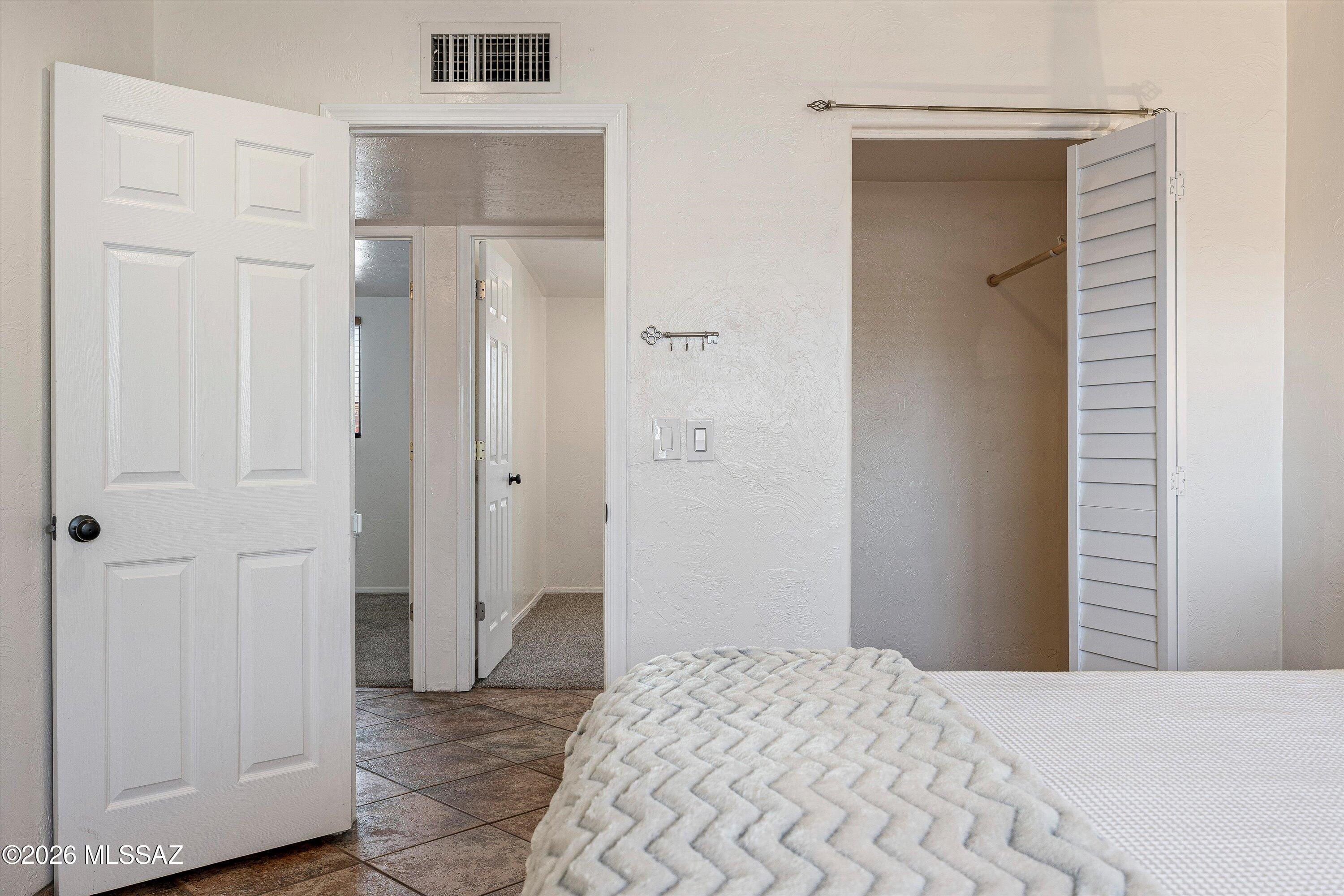 Bedroom with a white bed, open louvered closet, and tiled floor by the doorway.