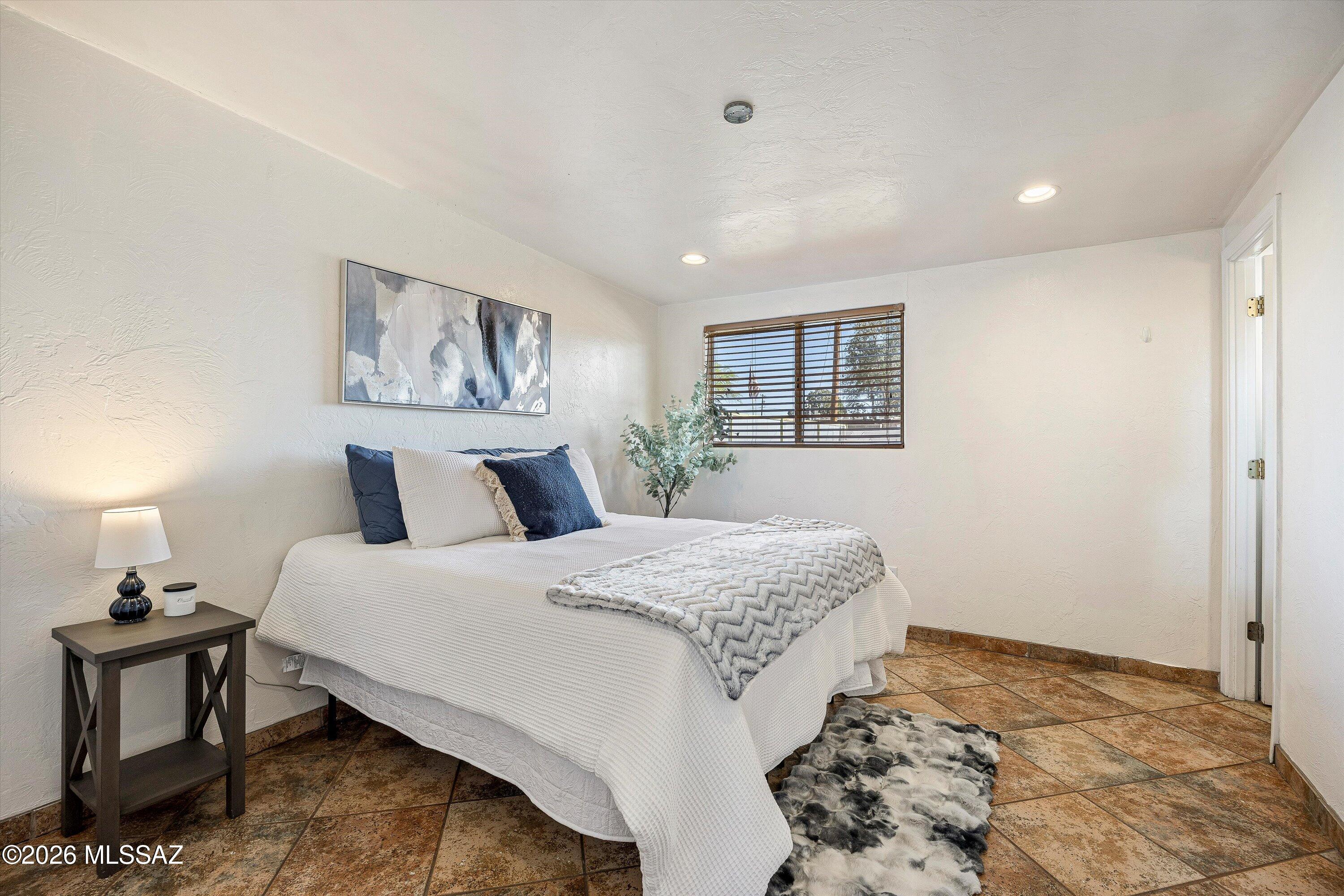 Staged bedroom with a white bed, patterned tile floor, and a window with wooden blinds.