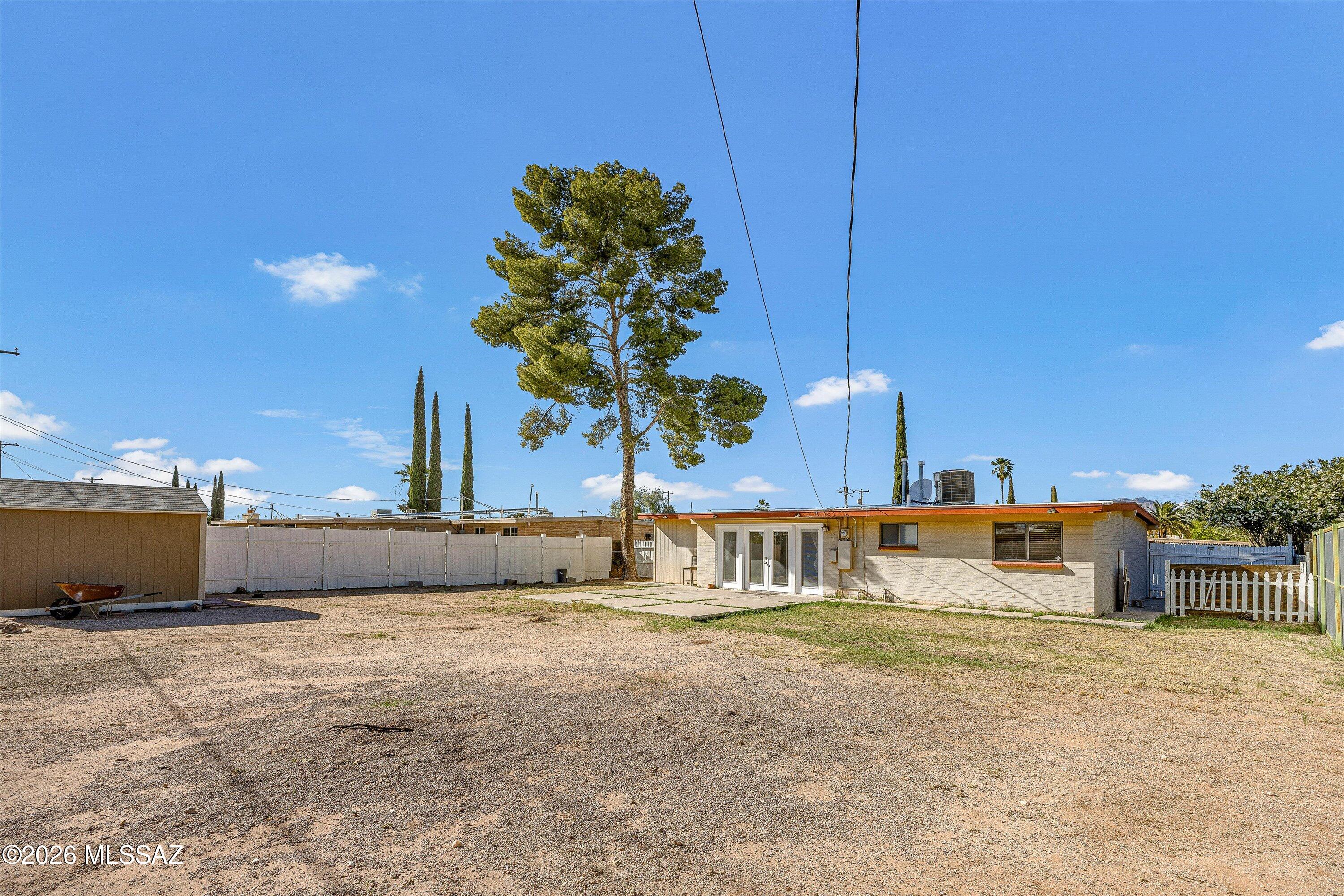 Single-story home viewed across a gravel yard, with a tall pine tree behind it.