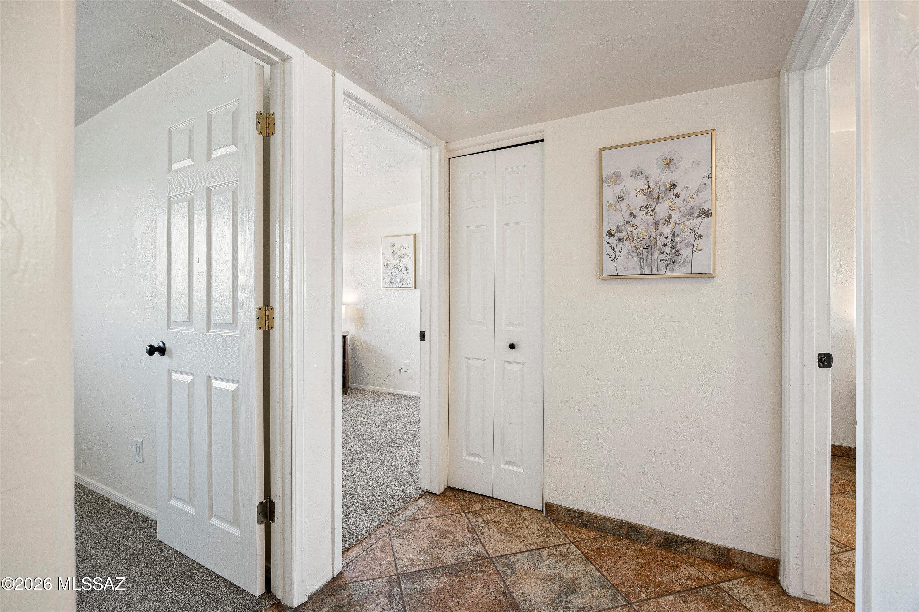 Hallway with tan tile flooring, white doors, a double-door closet, and framed floral artwork.