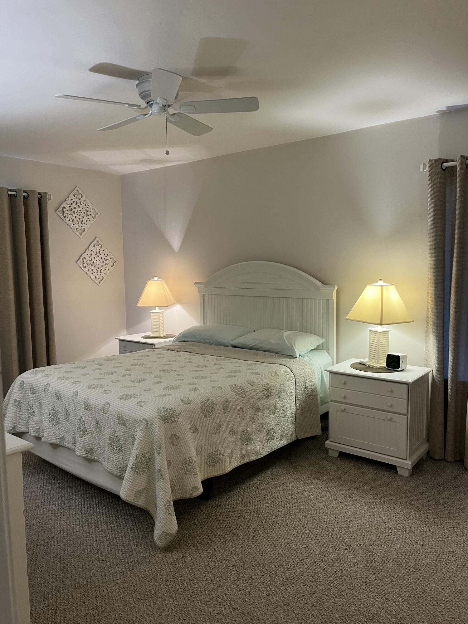 Furnished bedroom with a white bed, patterned quilt, matching nightstands, and ceiling fan.