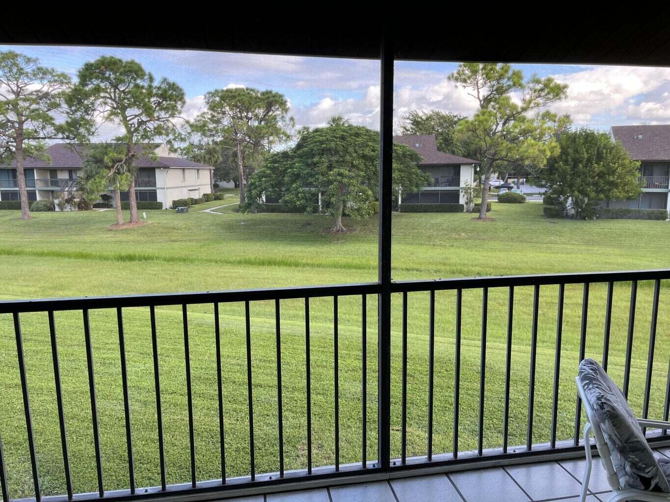 Condo balcony overlooking a broad lawn, mature trees, and neighboring residential buildings.