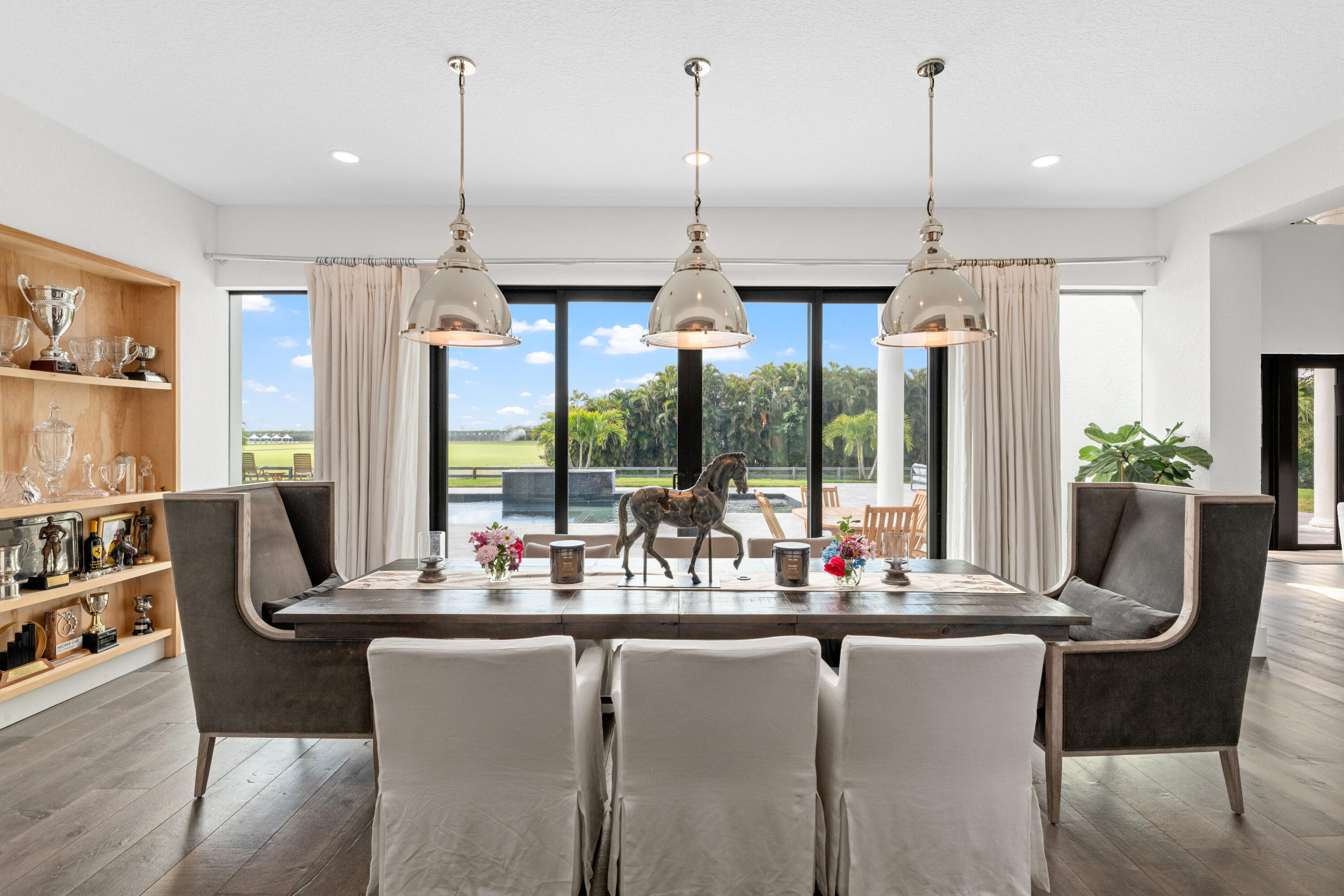 Dining room with a wood table, trophy shelves, and pool and palm views through sliding glass doors.